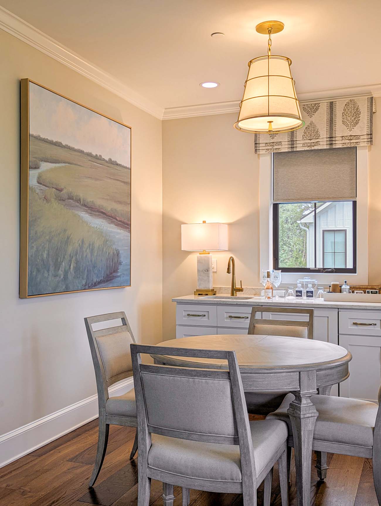 Intimate dining space with round gray wooden table surrounded by upholstered chairs, illuminated by a large nautical-style pendant light with brass accents. A prominent landscape painting depicting marshland and waterways dominates one wall, while windows with patterned valances provide natural light. Built-in cabinetry with sink and small appliances line the opposite wall.