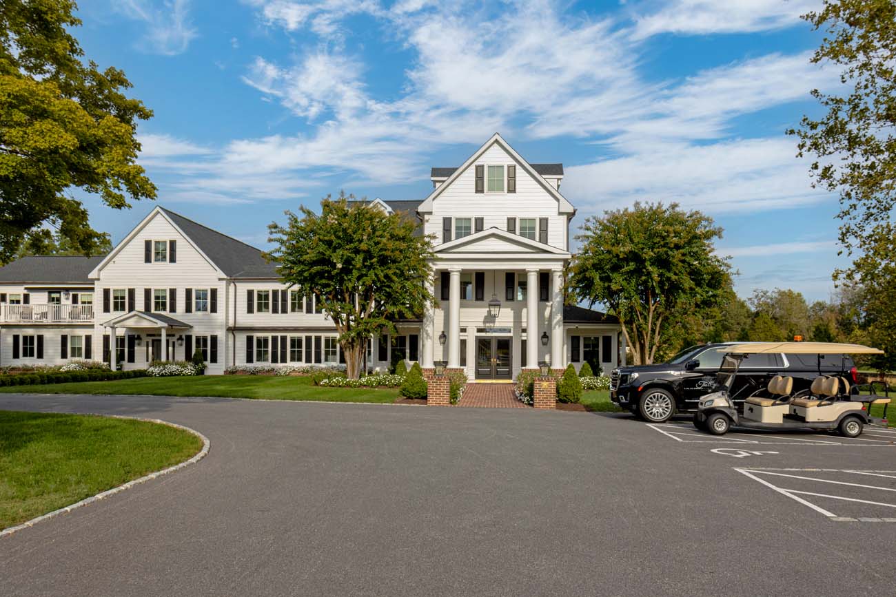 Impressive white colonial revival hotel building featuring multiple gables, black shutters, and covered entry portico supported by columns. The circular driveway is paved with dark asphalt and includes golf cart parking areas. Manicured landscaping with foundation plantings and a brick-edged entrance create an elegant arrival experience.