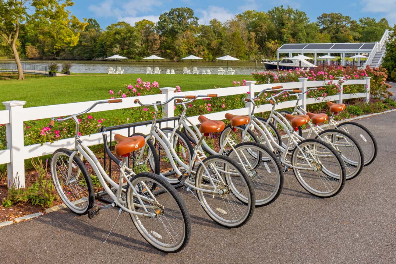 Six cream-colored cruiser bikes with brown leather seats lined up along white wooden fencing, with vibrant flower beds in foreground. Beyond lies expansive green lawn leading to waterfront with white Adirondack chairs under umbrellas, boat dock with covered pavilion, and luxury yacht moored at pier. Trees frame the scenic Chesapeake Bay setting.