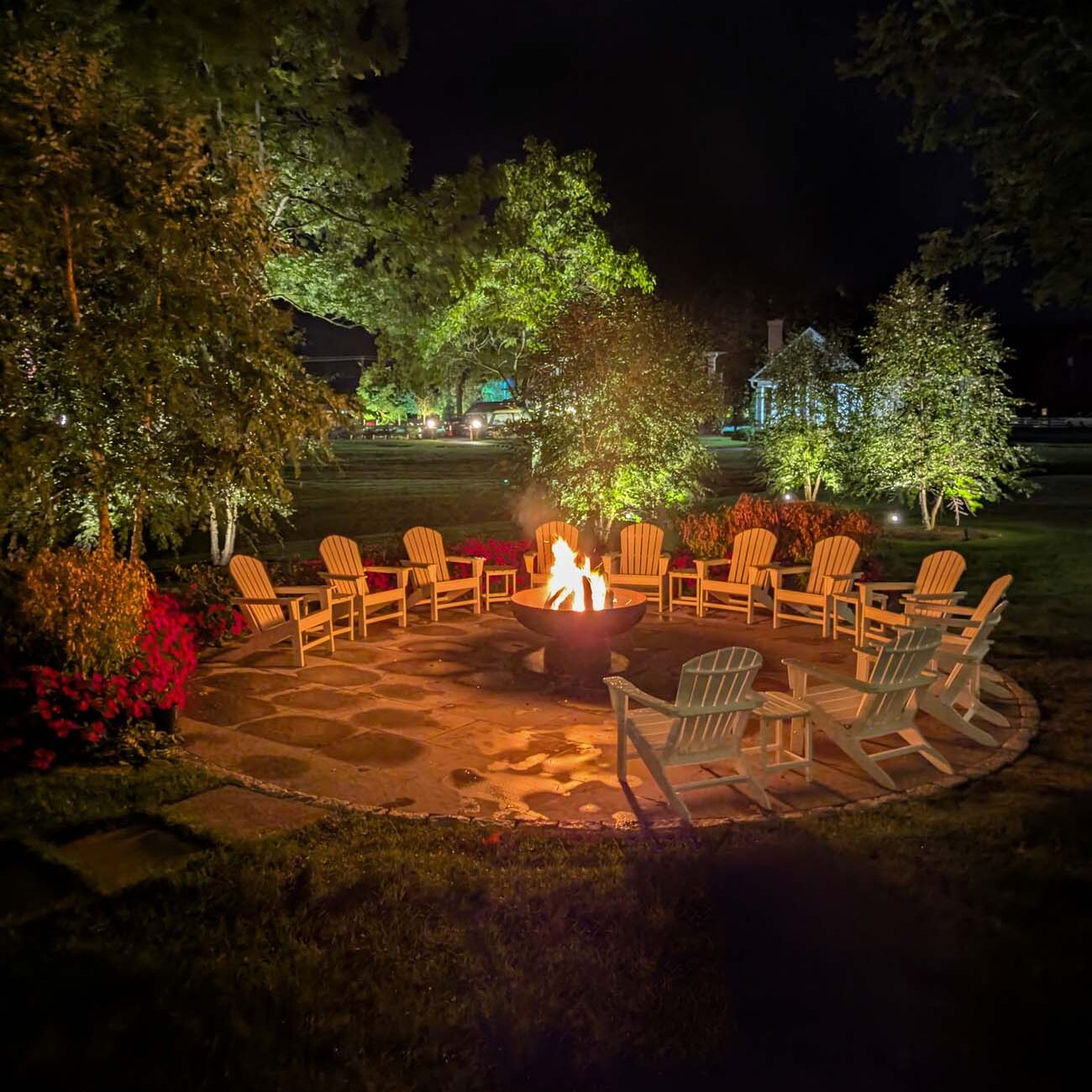  Circular stone fire pit surrounded by multiple wooden Adirondack chairs arranged on flagstone patio, with flames visible in the center. The scene is illuminated against dark evening sky, with mature trees and cottage lights creating ambient background lighting. Additional seating areas are visible in the distance, emphasizing the social nature of this outdoor gathering space.