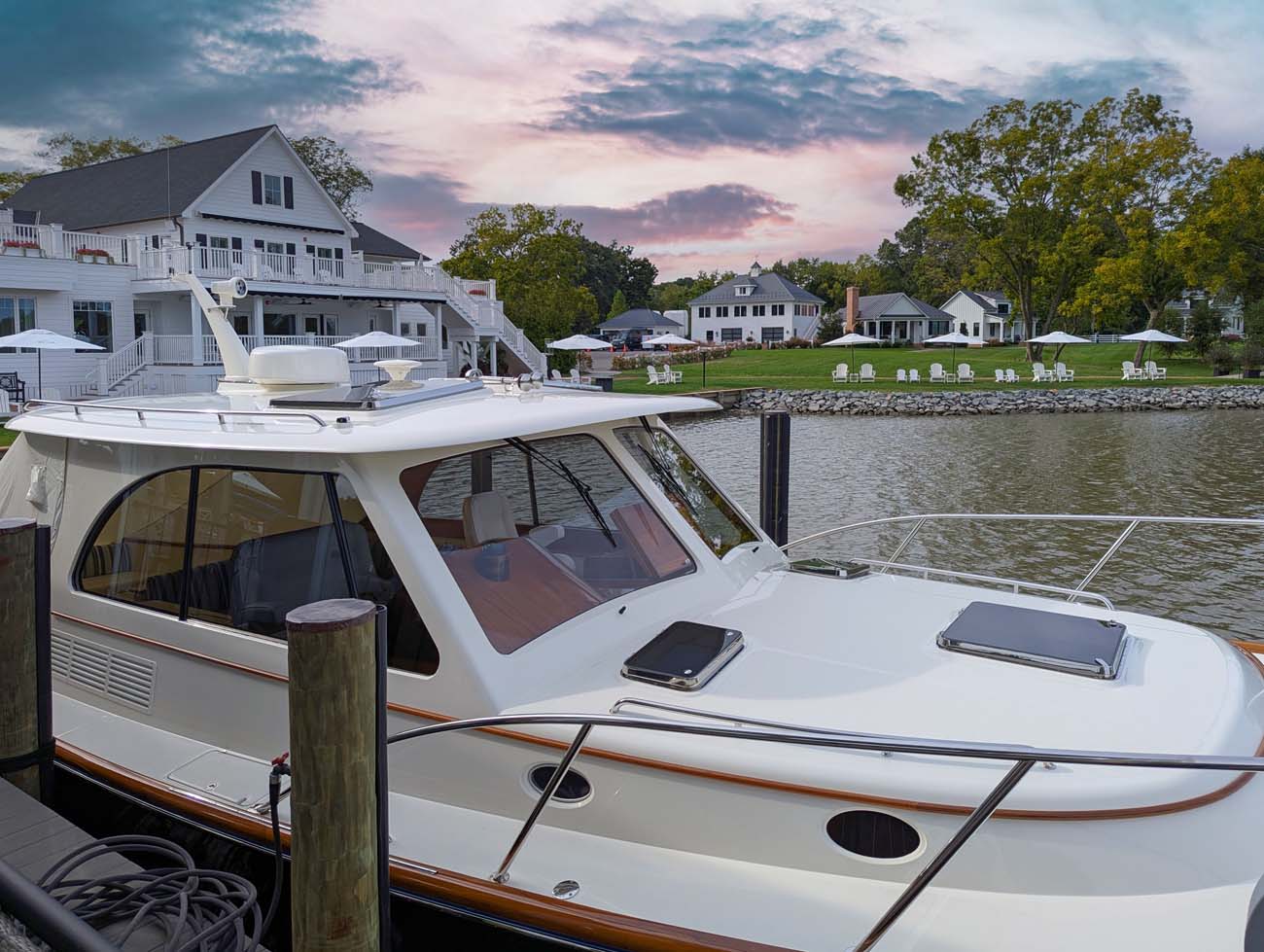 Luxury white yacht with enclosed cabin and dark trim moored at wooden dock during golden hour. The 40-foot vessel features multiple windows, professional rigging, and classic yacht styling. In the background, the hotel's white buildings with multiple levels and balconies are visible across the manicured waterfront lawn dotted with white outdoor furniture.