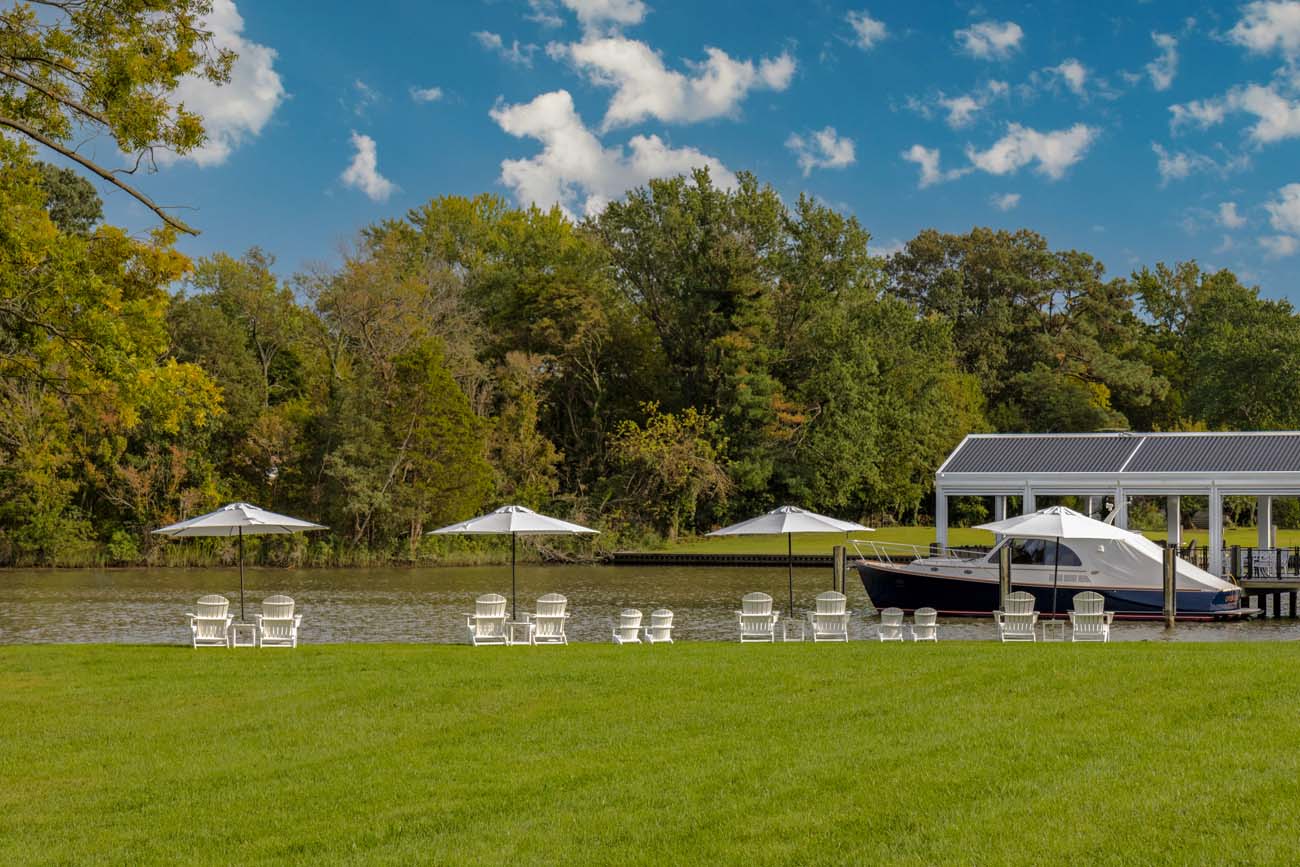 Expansive green lawn stretching to waterfront with multiple groupings of white Adirondack chairs positioned under beige umbrellas for sun protection. A covered boat slip houses a dark-hulled yacht, while mature trees provide natural backdrop. The peaceful setting emphasizes relaxation and water access amenities.