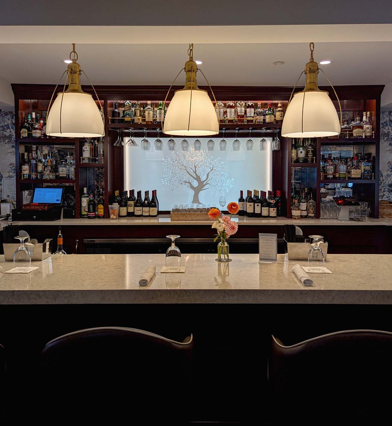 Elegant hotel bar with rich dark wood cabinetry, marble countertop, and three large nautical-style pendant lights with cream shades and brass hardware. Wine bottles line built-in shelving, wine glasses hang from overhead racks, and decorative tree motif provides artistic backdrop. Two modern bar stools with curved backs face the professionally equipped bar station.