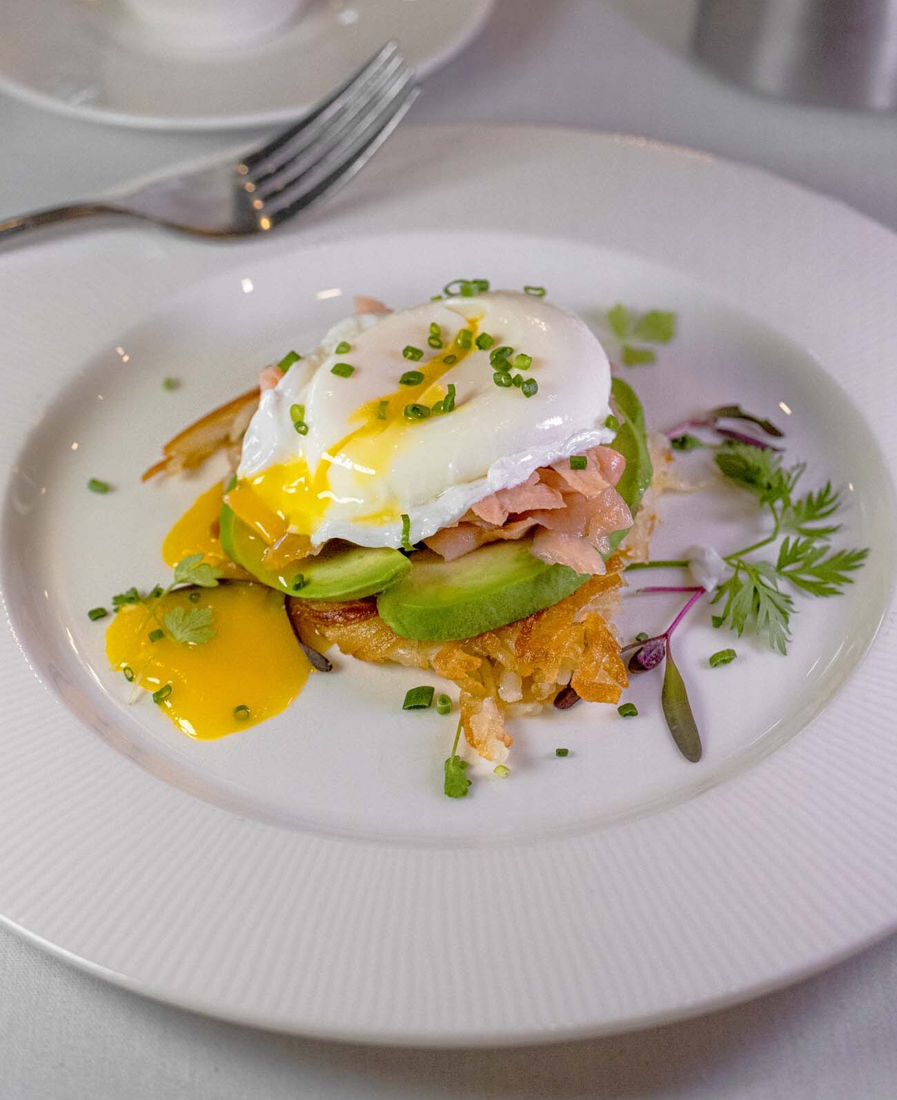 Artfully plated breakfast dish featuring perfectly poached egg with runny golden yolk atop rösti potatoes, smoked salmon, and sliced avocado. The white ceramic plate is garnished with microgreens and colorful edible flowers, with bright yellow yolk creating dramatic color contrast. Professional fine dining presentation demonstrates culinary attention to detail.