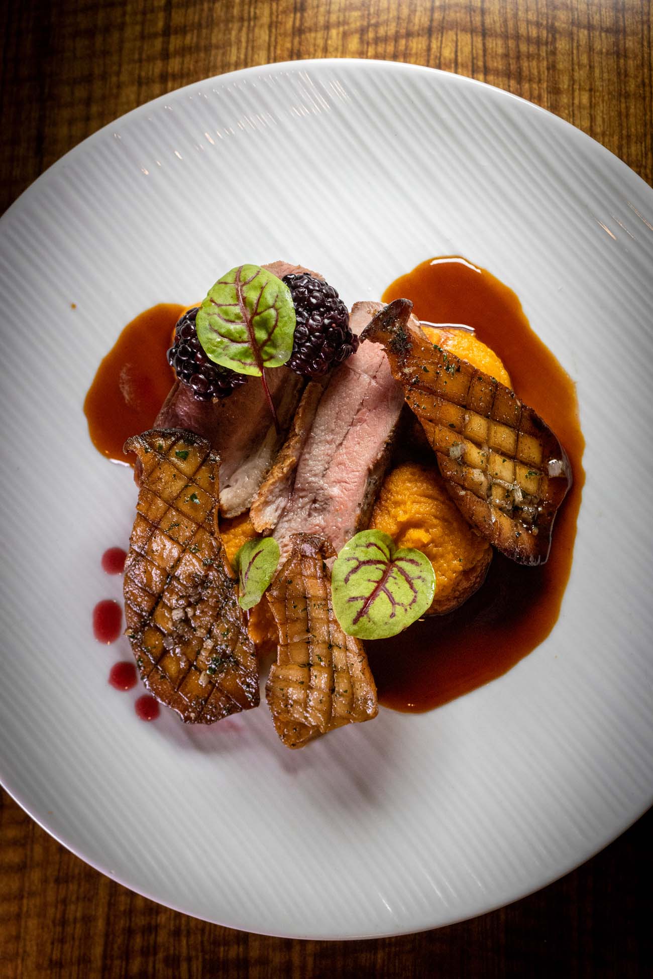 Sophisticated dinner plate featuring sliced duck breast cooked to perfect medium-rare with pink interior, accompanied by bright orange sweet potato puree, seared royal trumpet mushrooms with crosshatch scoring, pickled blackberries, and decorative microgreens. Rich brown port wine reduction sauce pools around the elements on the pristine white ceramic plate.