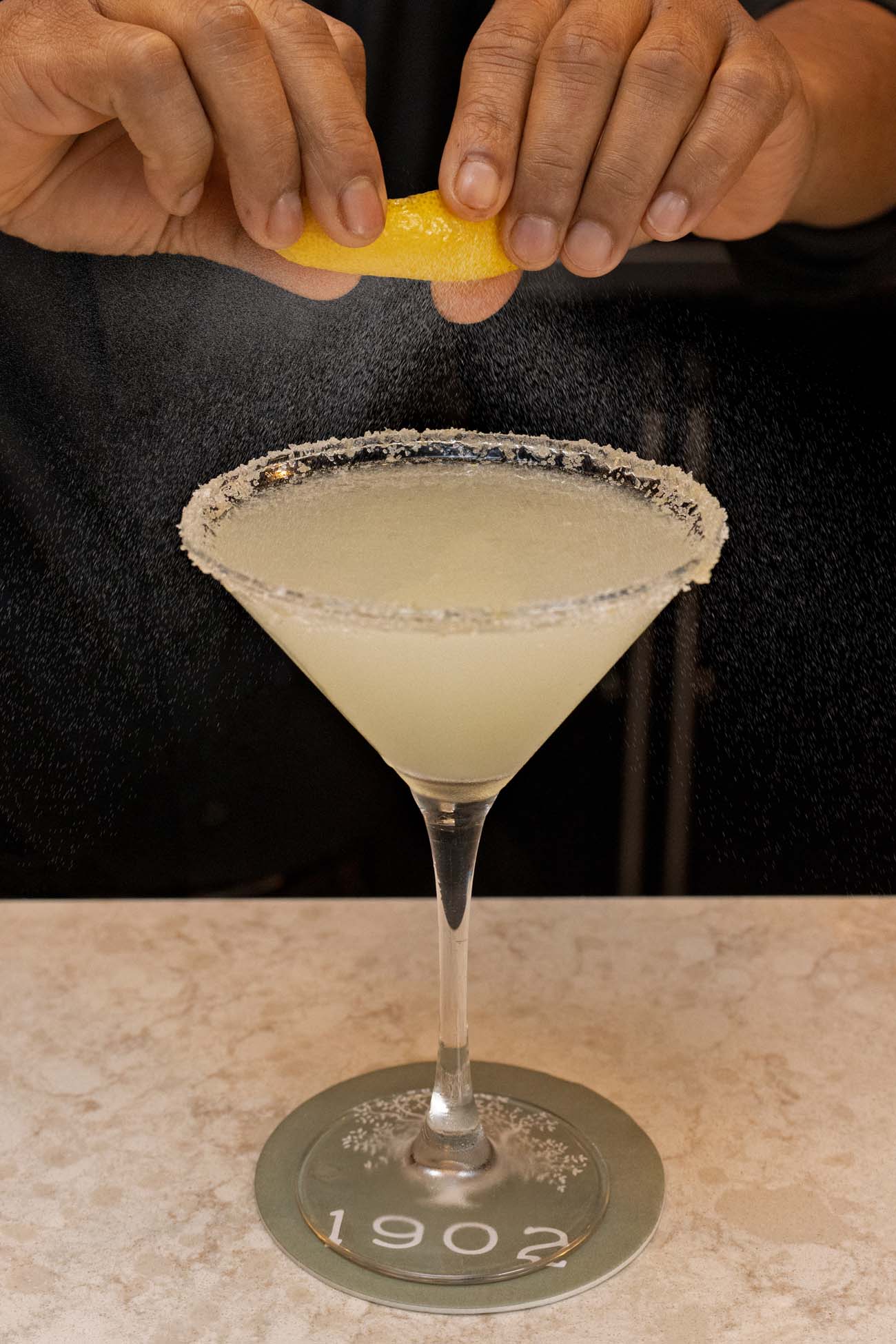 Hands squeezing fresh lemon peel over a pale yellow cocktail in a martini glass with salted rim. The citrus oils are visibly misting as they're expressed from the bright yellow lemon peel. The drink sits on a distinctive coaster marked "1902" referencing the hotel's founding year, positioned on a marble bar surface against a dark background.
