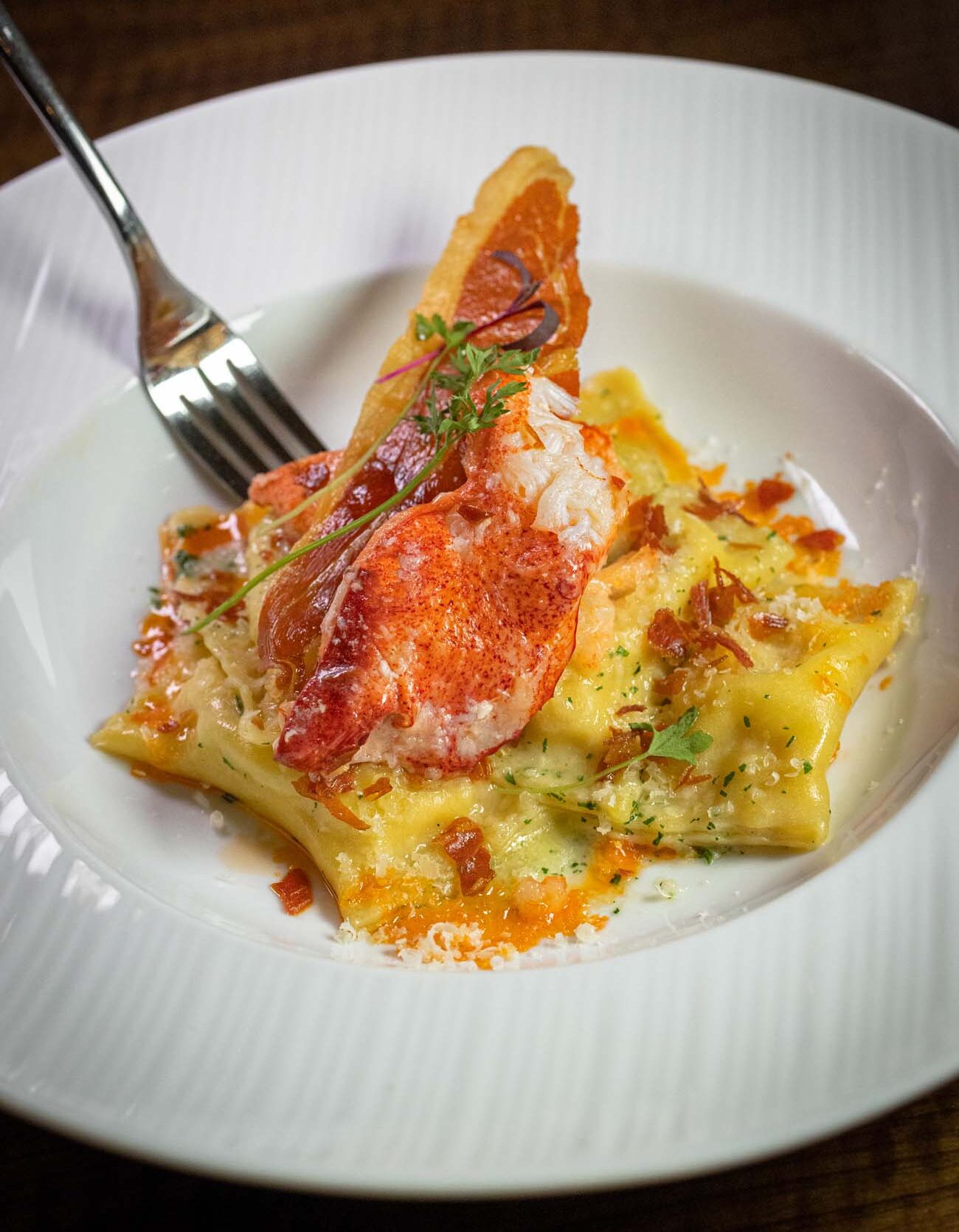 Elegant white ceramic plate featuring house-made pasta topped with chunks of fresh lobster meat, crispy golden pancetta pieces, and fresh herbs. A silver fork rests beside the dish, and the pasta appears to be in a light sauce with visible herbs and pancetta creating textural contrast against the tender lobster and pasta.