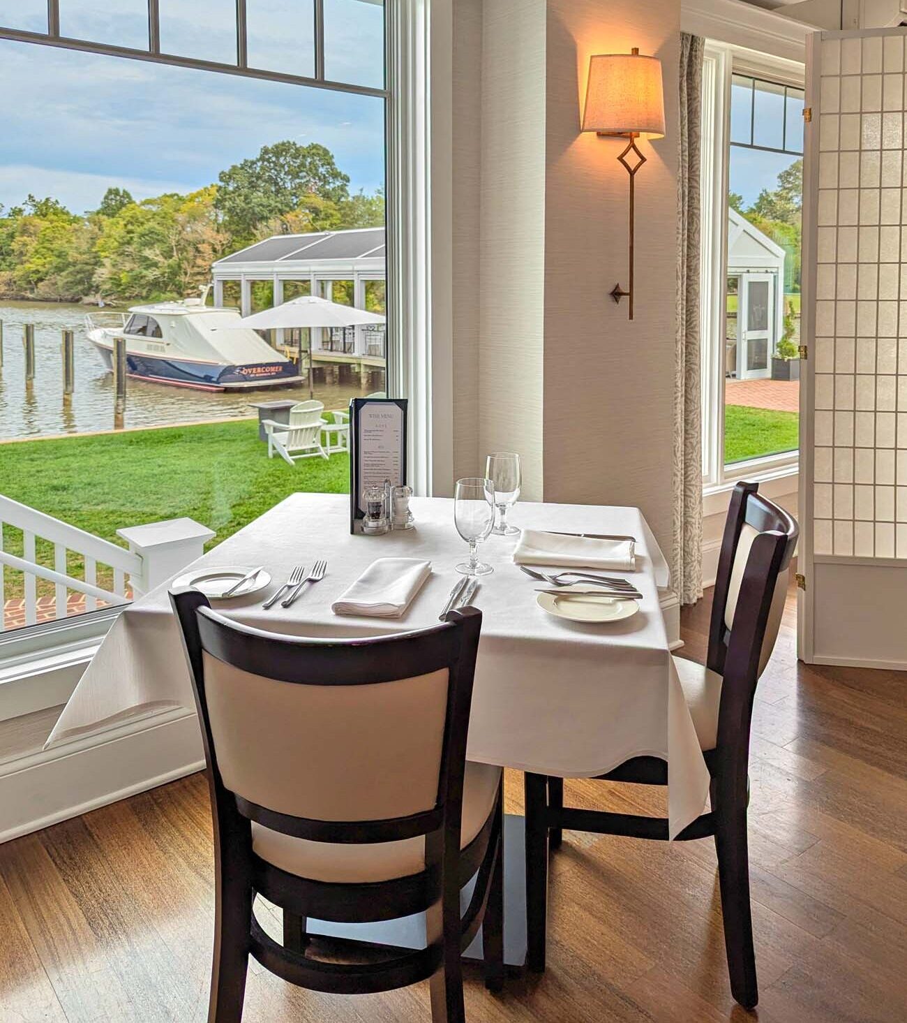 Intimate two-person dining table set with white linens, wine glasses, and formal place settings positioned beside large windows overlooking Oak Creek. The "Overcomer" yacht is visible at the dock, while warm evening light filters through the space. Professional table setting and scenic water views create an elegant fine dining atmosphere.