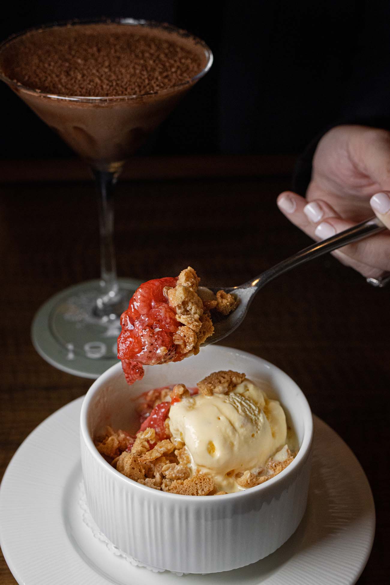 White ceramic ramekin containing strawberry crisp dessert topped with vanilla ice cream and garnished with fresh strawberries and crumble topping. A spoon holds a bite of the dessert showing the layered textures. In the background, a chocolate martini with rich brown color and chocolate rim garnish sits on the signature "1902" coaster.