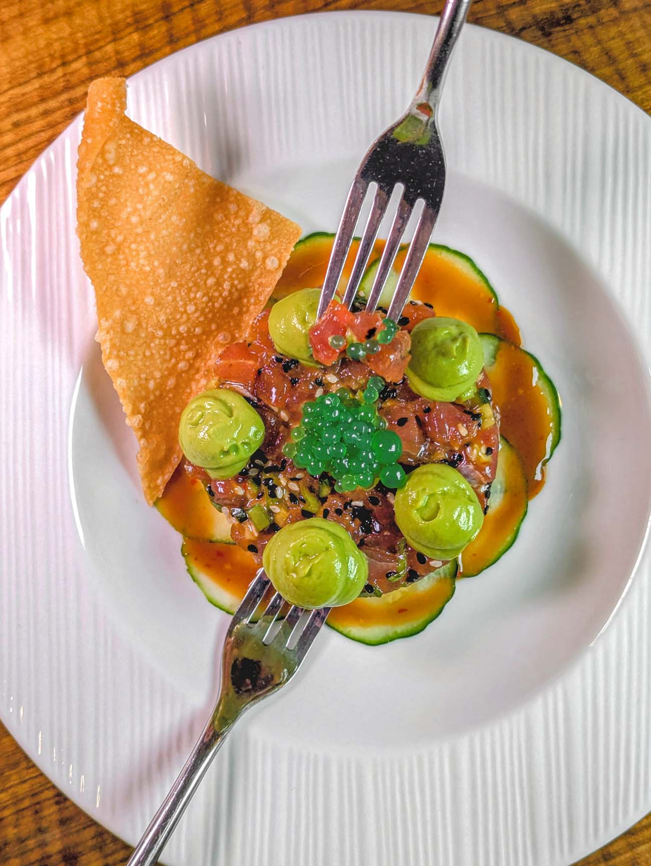 Artfully plated tuna tartare on white ceramic plate featuring diced raw tuna mixed with sesame seeds, lime, and tamari, topped with bright green caviar and sliced cuvumbers. The dish is accompanied by cucumber ribbons, avocado mousse dollops, and a golden crispy wonton chip. Two forks frame the colorful presentation on a warm wood table surface.