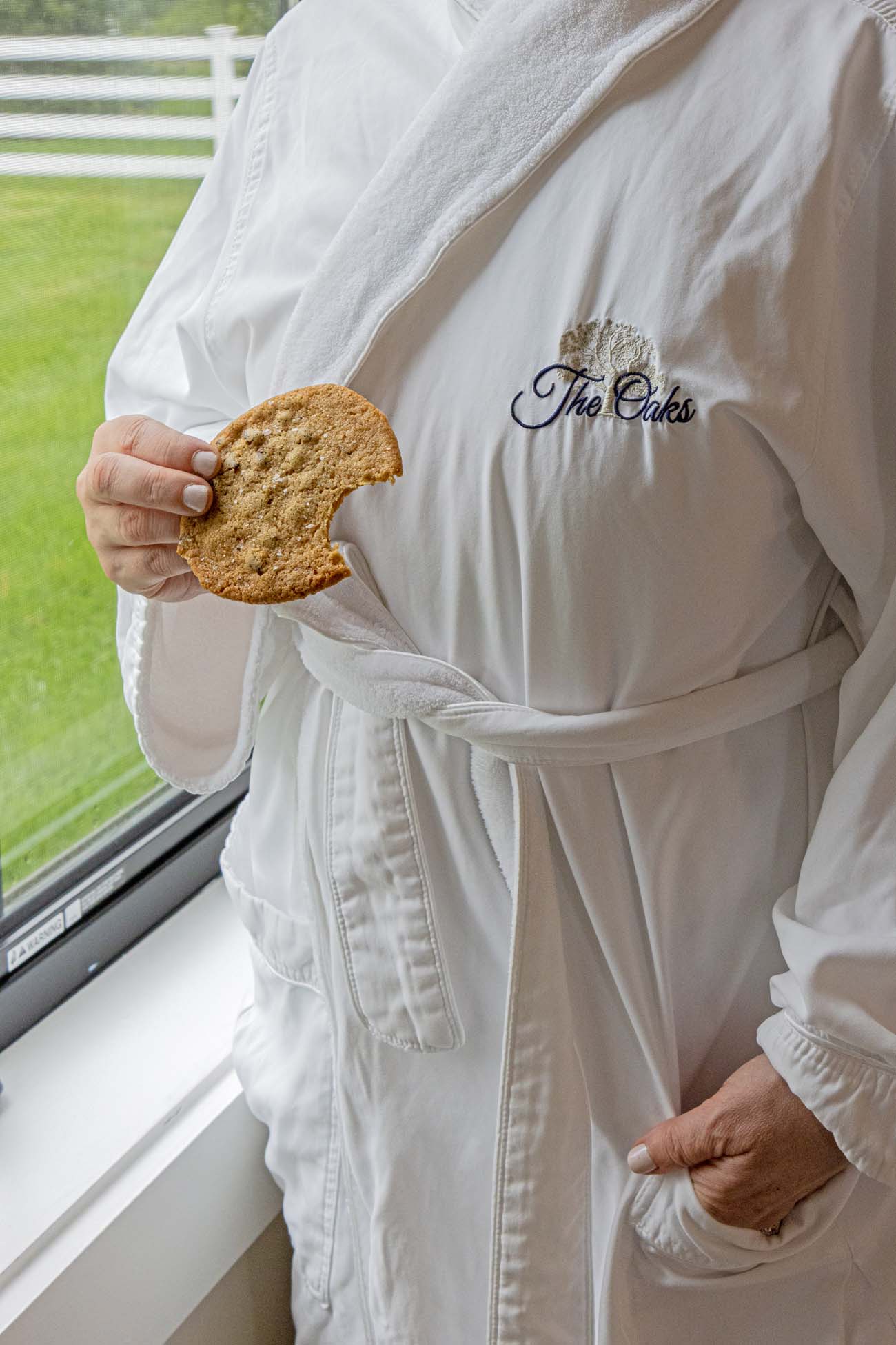 Person wearing plush white terrycloth bathrobe embroidered with "The Oaks" logo and decorative tree design, holding a partially eaten chocolate chip cookie while standing by large windows overlooking green grounds with white fencing. The scene captures the relaxed, pampered atmosphere of the waterfront hotel experience.