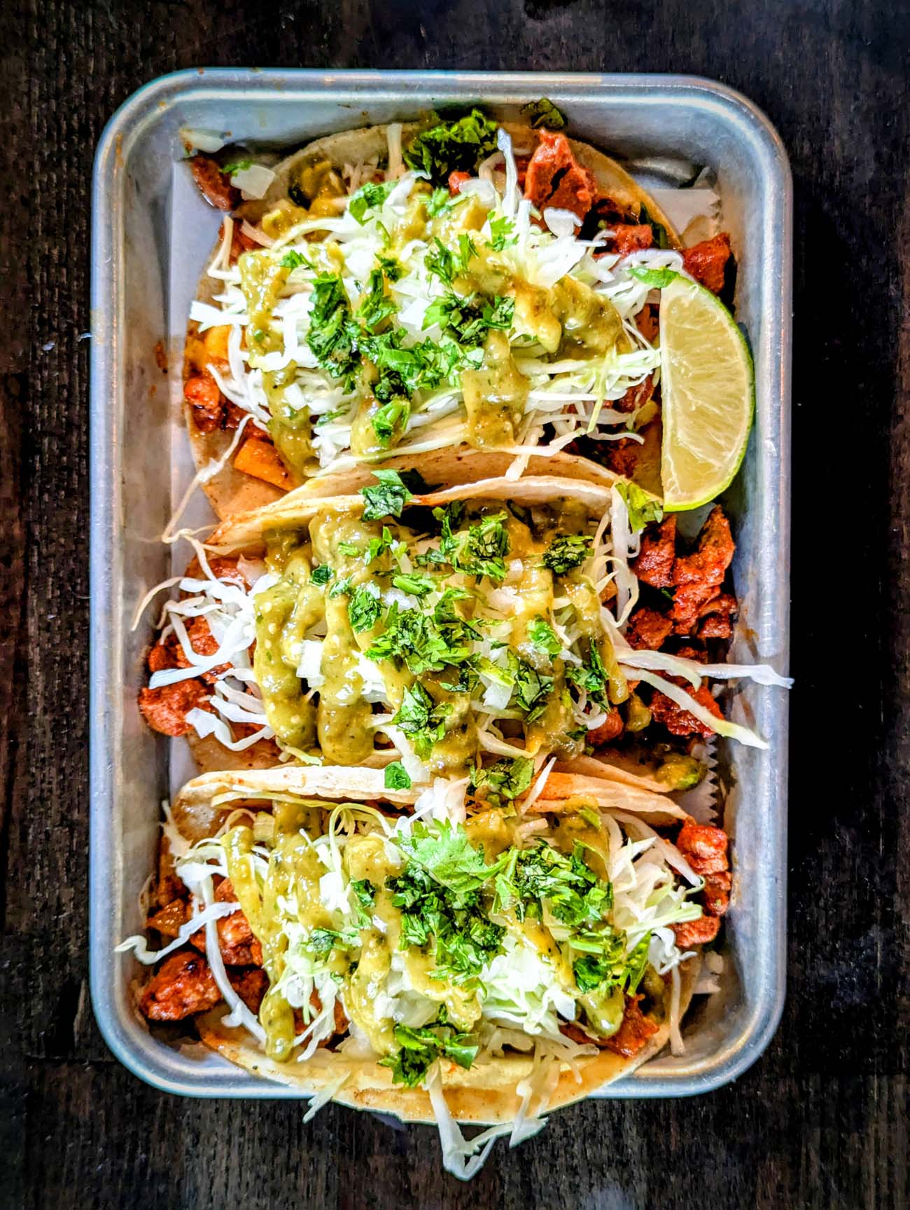  Silver aluminum takeout container holding three soft tacos filled with adobo-marinated pork, topped with fresh cilantro, white shredded cheese, and green salsa verde. A lime wedge provides citrus accent, and the tacos are garnished with additional herbs and appear to include pineapple pieces characteristic of al pastor preparation.
