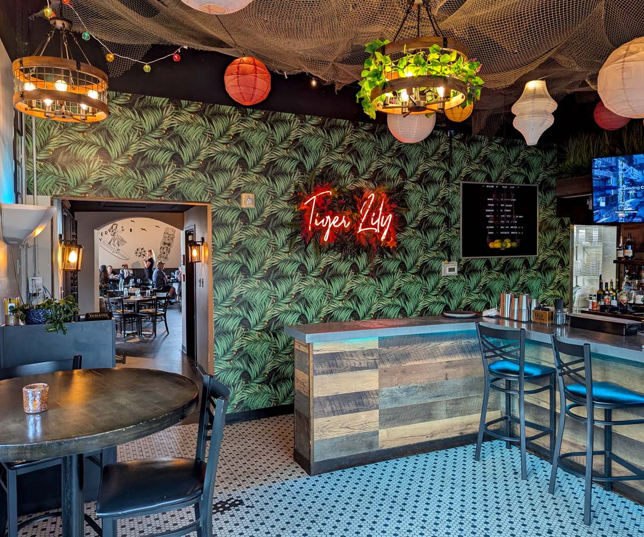 Vibrant restaurant interior featuring dark green palm leaf wallpaper, fishing net ceiling decoration with colorful paper lanterns, and red neon "Tiger Lily" sign as focal point. The space includes reclaimed wood bar seating, round tables, and eclectic lighting creating a fun, tropical atmosphere. An open kitchen and dining room are visible in the background, with string lights and tiki-inspired design elements throughout.