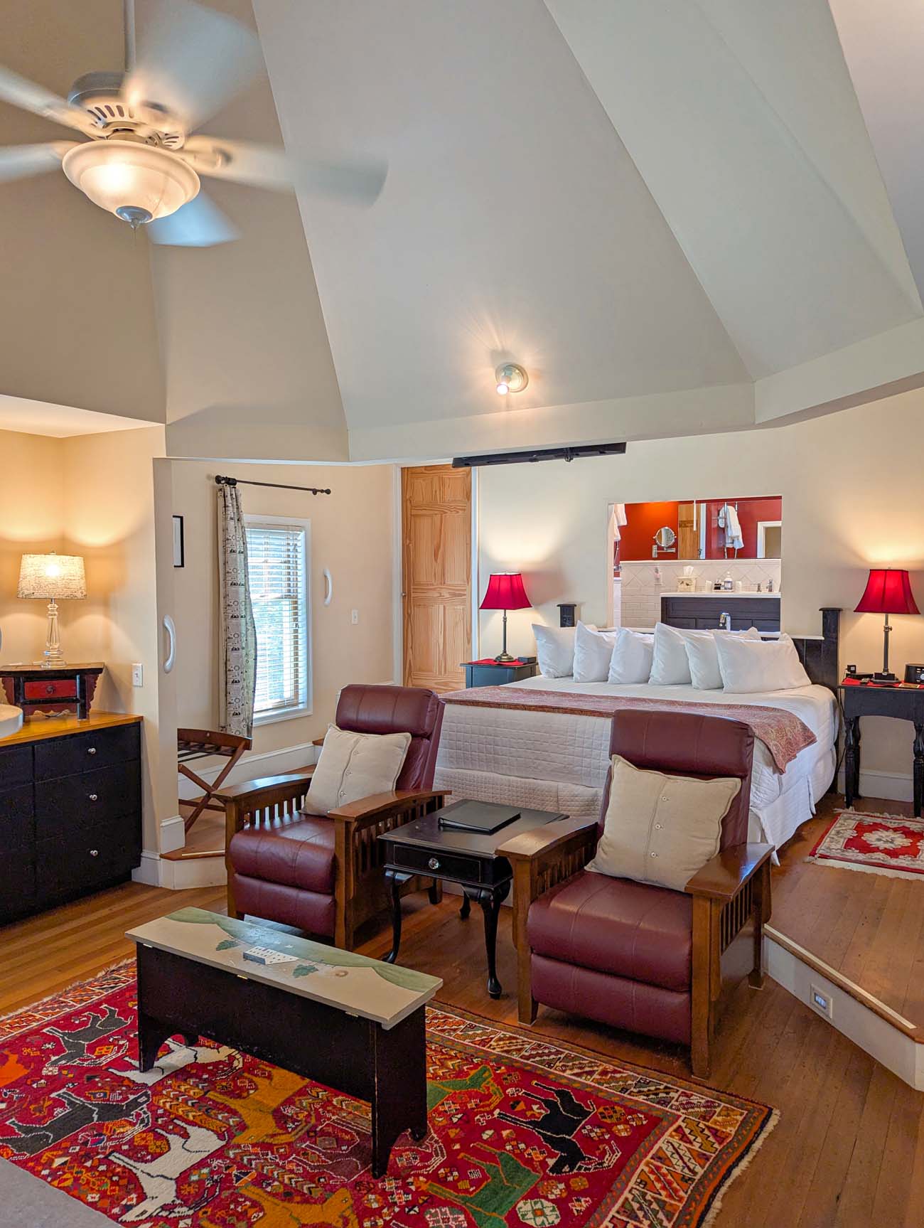 Food, Wine, and Fall Fun at Brampton 1860 - Chestertown, MD 6 Bright, airy cottage bedroom with vaulted ceiling, king bed with white linens and burgundy leather bench at foot, two burgundy leather recliners, sliding barn door to bathroom, red accent lamps, colorful area rug, hardwood floors