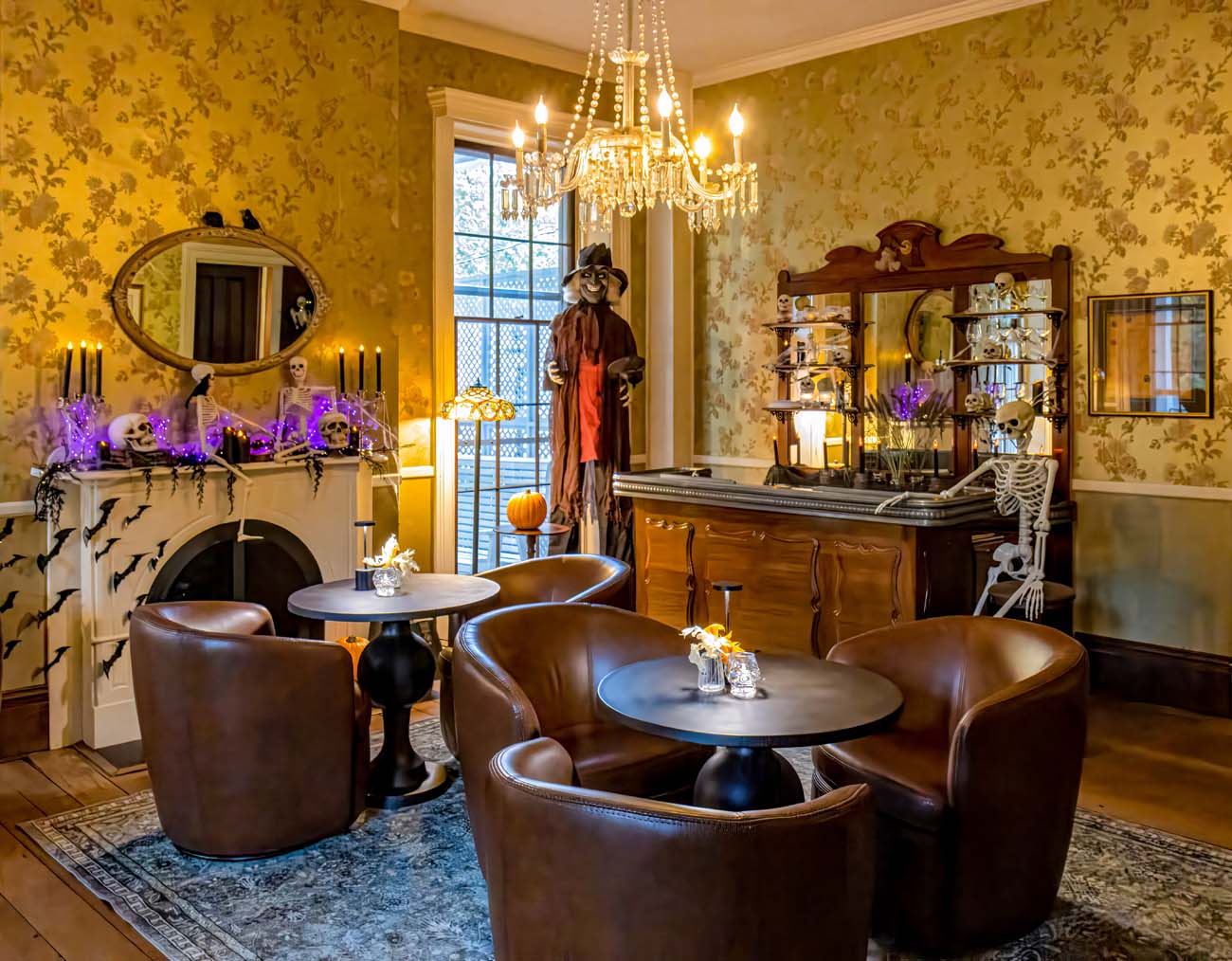 Food, Wine, and Fall Fun at Brampton 1860 - Chestertown, MD 36 Elegant Manor House lounge transformed for Halloween with antique wooden bar, skeleton bartender prop, life-size witch figure in Victorian dress, crystal chandelier, gold wallpaper, fireplace decorated with black paper bats, purple LED lights, black candles on mantel, leather club chairs around small tables, oriental rug on hardwood floors
