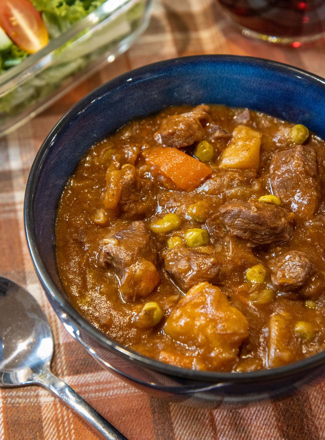 A rich bowl of hearty beef stew with a spoon next to a salad and glass of wine.