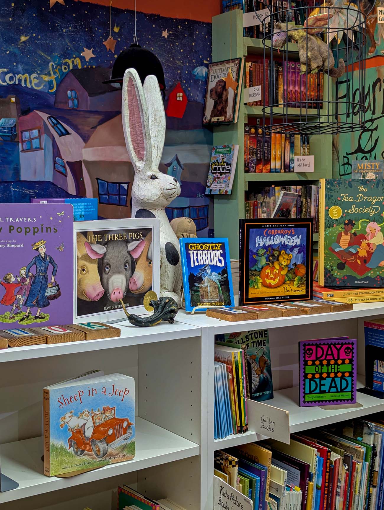 Things to Do in Chestertown MD: Eastern Shore Travel Guide 16 Interior of The Bookplate independent bookstore in Chestertown Maryland showing white bookshelves filled with children's books including The Three Pigs, Ghostly Terrors, and Halloween books, with a large white decorative rabbit statue, whimsical wall mural with stars and houses in background