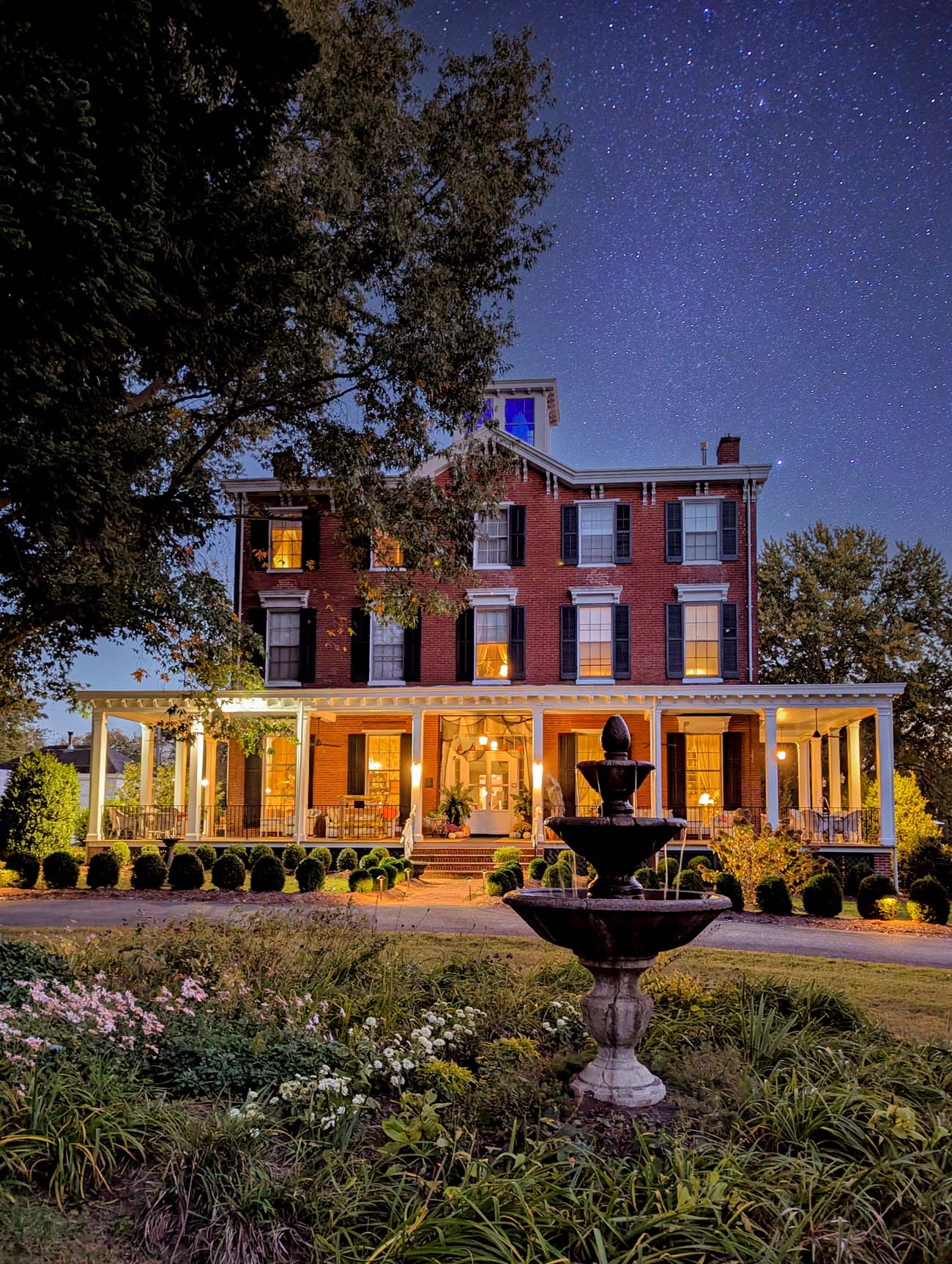 Things to Do in Chestertown MD: Eastern Shore Travel Guide 30 Brampton 1860 historic brick inn at dusk with starry night sky, showing three-story red brick building with white wraparound porch, glowing windows, decorative fountain in manicured garden with colorful flowers, trees on either side