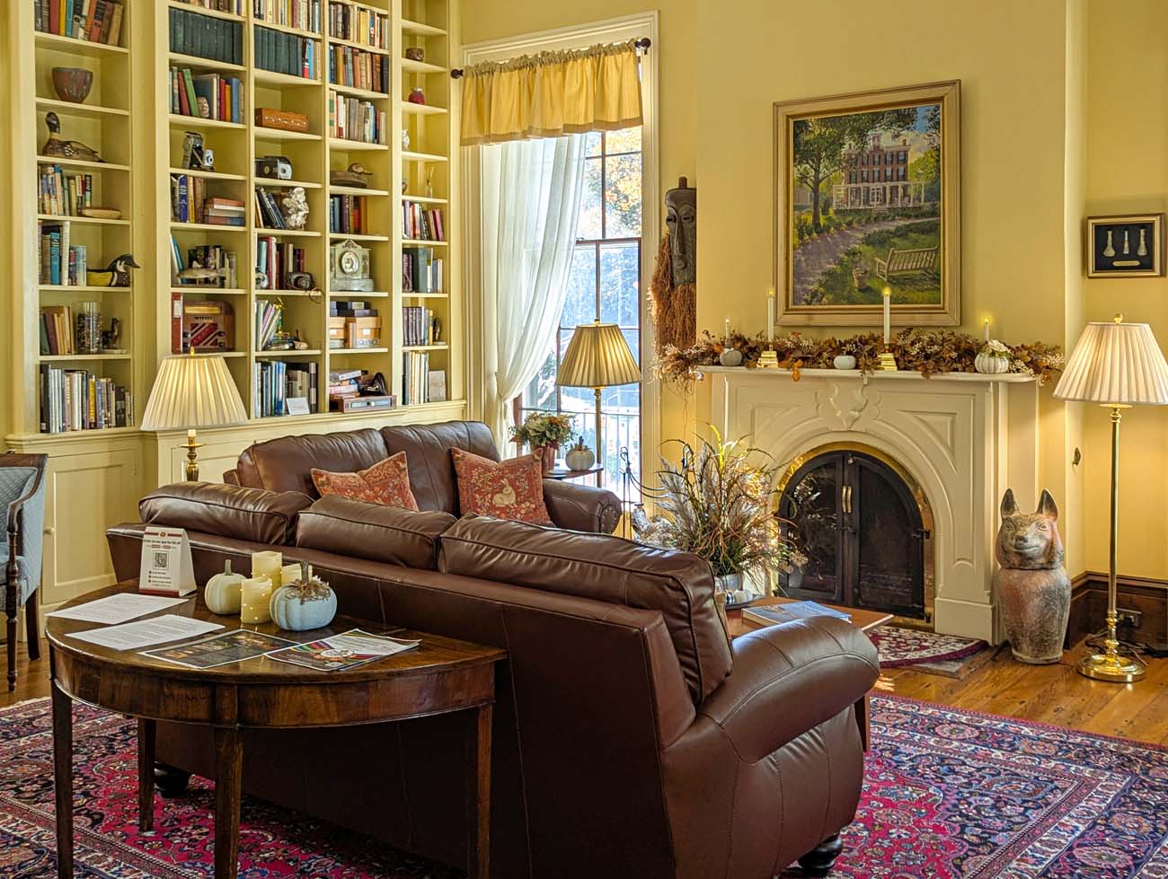 Things to Do in Chestertown MD: Eastern Shore Travel Guide 36 Cozy library room at Brampton 1860 with floor-to-ceiling white built-in bookshelves, brown leather sofa with rust-colored pillows, arched fireplace with white mantel decorated with fall pumpkins and dried flowers, oriental rug, table lamps, and large window with yellow curtains