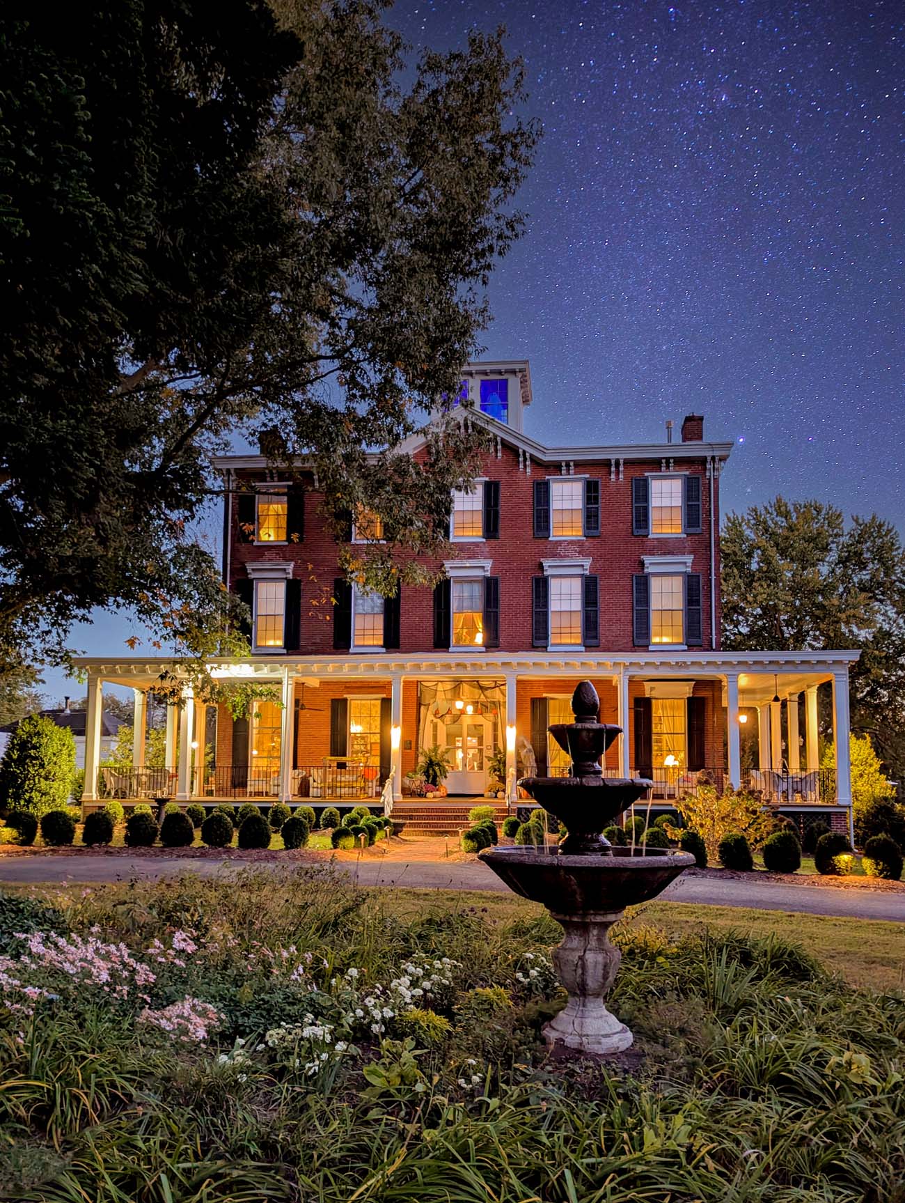 Brampton 1860 historic brick inn at dusk with starry night sky, showing three-story red brick building with white wraparound porch, glowing windows, decorative fountain in manicured garden with colorful flowers, trees on either side