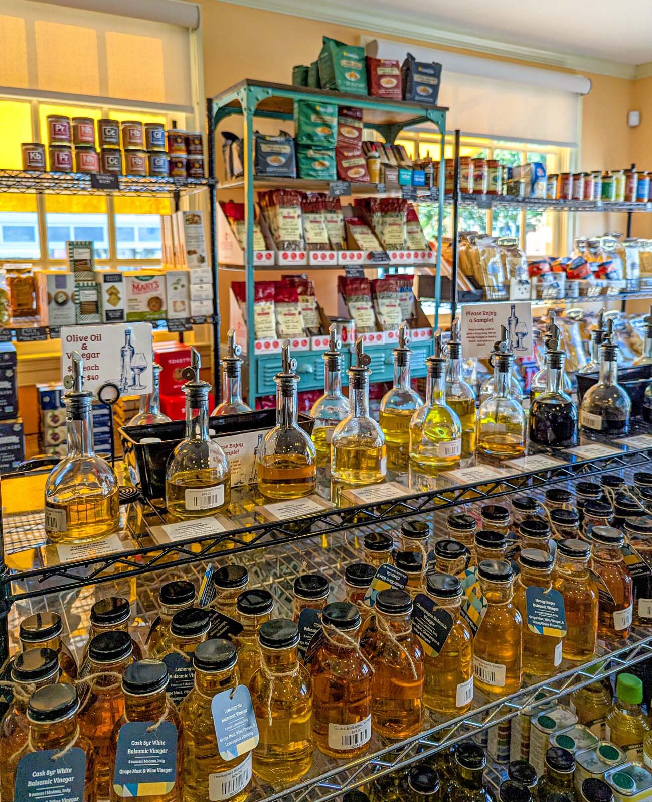 Things to Do in Chestertown MD: Eastern Shore Travel Guide 14 Interior of Chester River Wine & Cheese Co showing wire shelving filled with gourmet products including colorful pasta packages and specialty items on a turquoise shelf unit. In the foreground, a black wire rack displays multiple glass bottles of infused olive oils with pour spouts for sampling, alongside rows of small jars containing flavored honey in golden and amber hues. Additional shelves in the background hold various gourmet food products. Warm yellow walls and natural light from windows create an inviting atmosphere