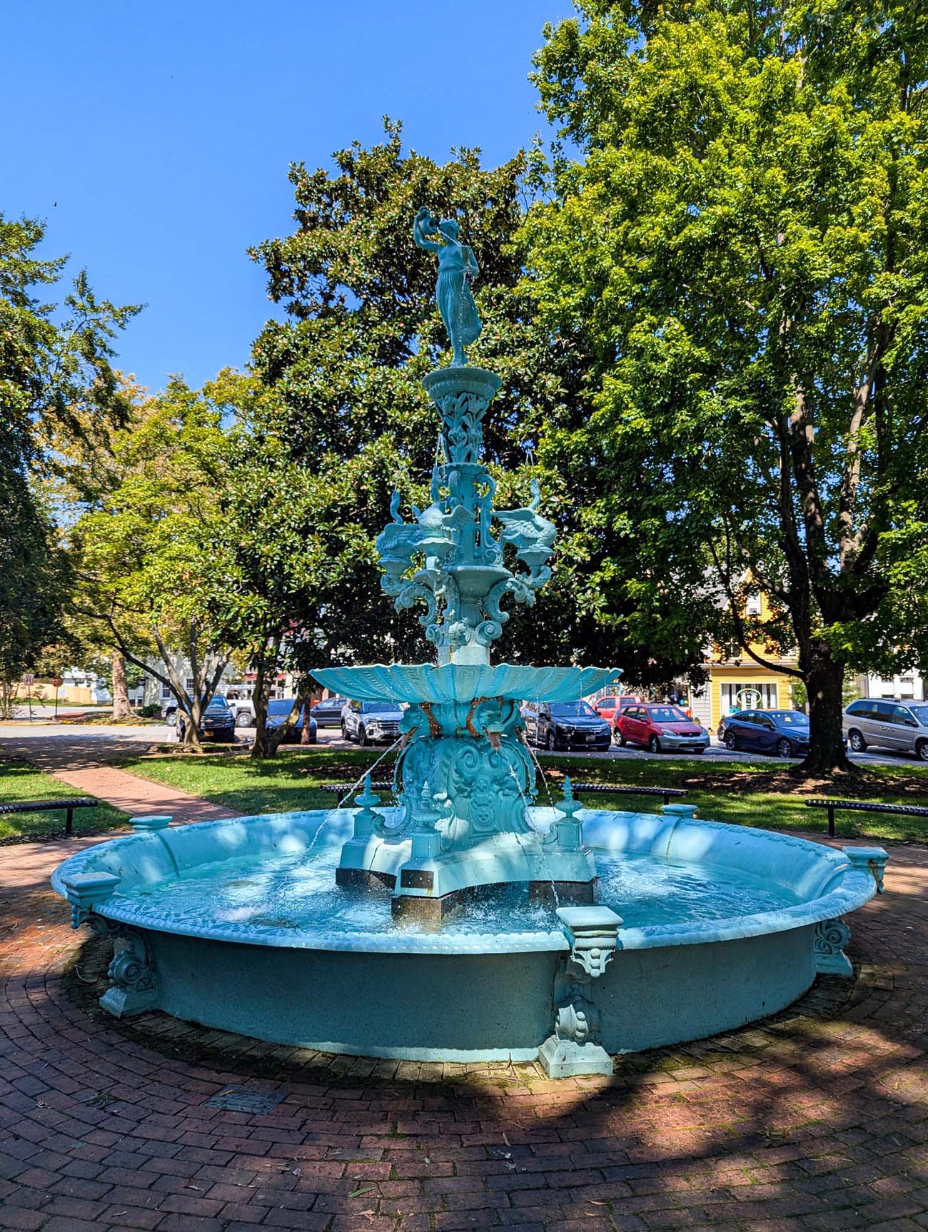 Things to Do in Chestertown MD: Eastern Shore Travel Guide 2 Ornate Victorian-era fountain painted in turquoise blue located in a town square in Chestertown, Maryland. The multi-tiered cast iron fountain features elaborate scrollwork, decorative flourishes, and a female figure statue at the top. Water flows from multiple levels into a circular basin below. The fountain sits on brick pavement surrounded by a green lawn, with mature trees providing dappled shade. Historic buildings and parked cars are visible in the background under a clear blue sky.