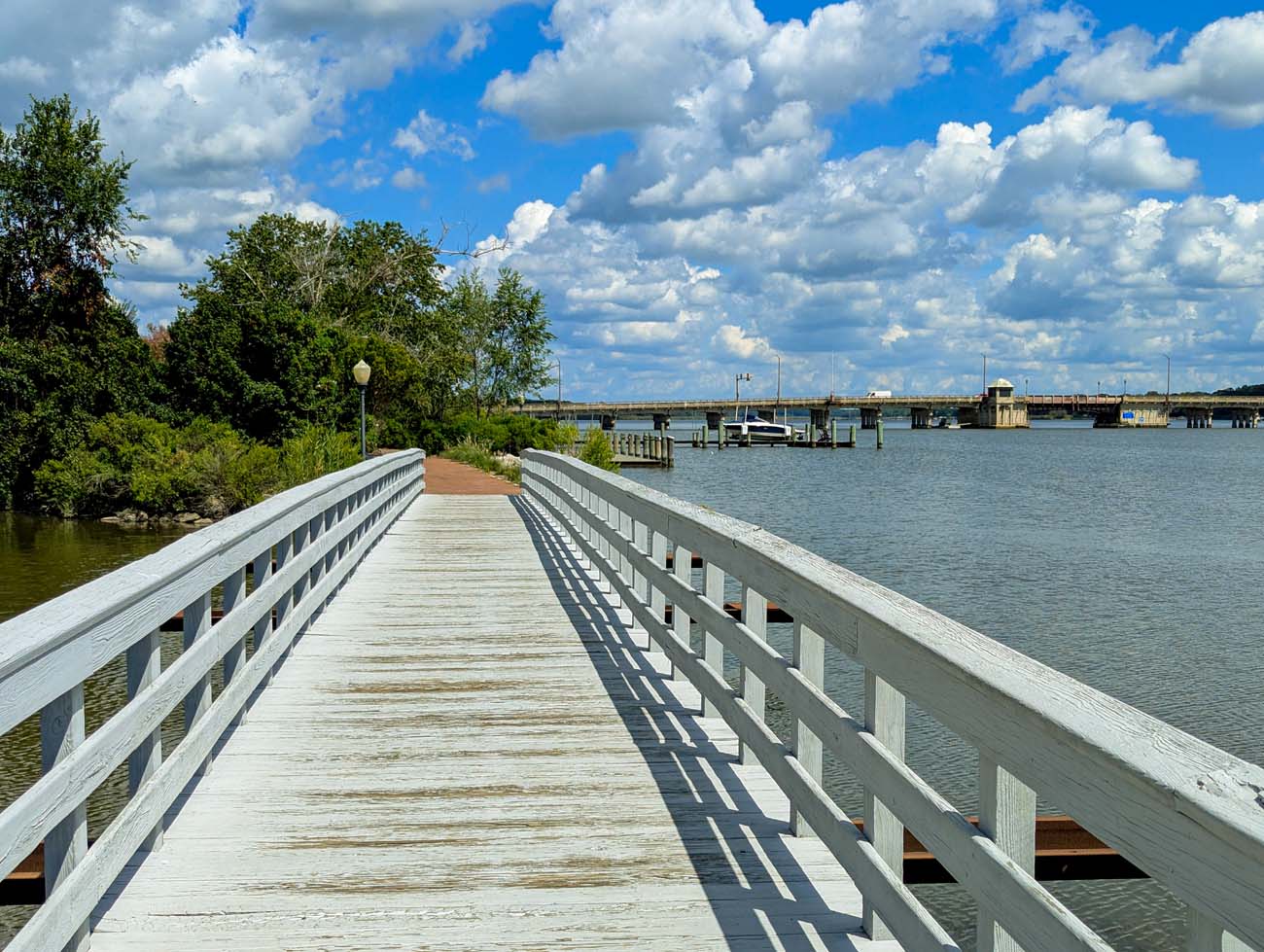 Things to Do in Chestertown MD: Eastern Shore Travel Guide 4 White wooden boardwalk pier extending over Chester River with gray railings on both sides, blue sky with white clouds, marina dock with boats visible in background, trees lining waterfront, peaceful water scene