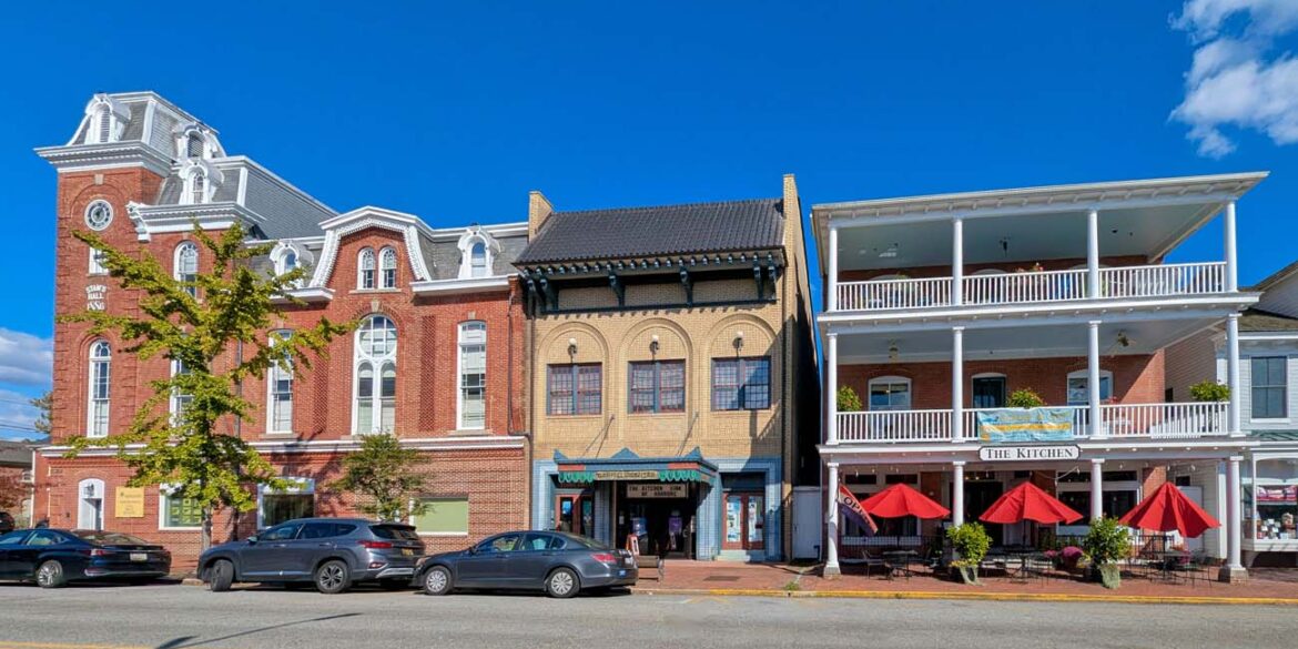 Historic downtown Chestertown Maryland street view showing row of preserved colonial and Victorian buildings including red brick Second Empire style town hall with clock tower, beige commercial building, and white building with double porches housing The Kitchen restaurant with red umbrellas, cars parked along street
