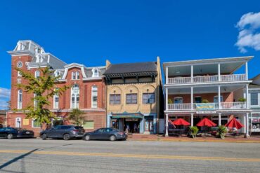 Historic downtown Chestertown Maryland street view showing row of preserved colonial and Victorian buildings including red brick Second Empire style town hall with clock tower, beige commercial building, and white building with double porches housing The Kitchen restaurant with red umbrellas, cars parked along street