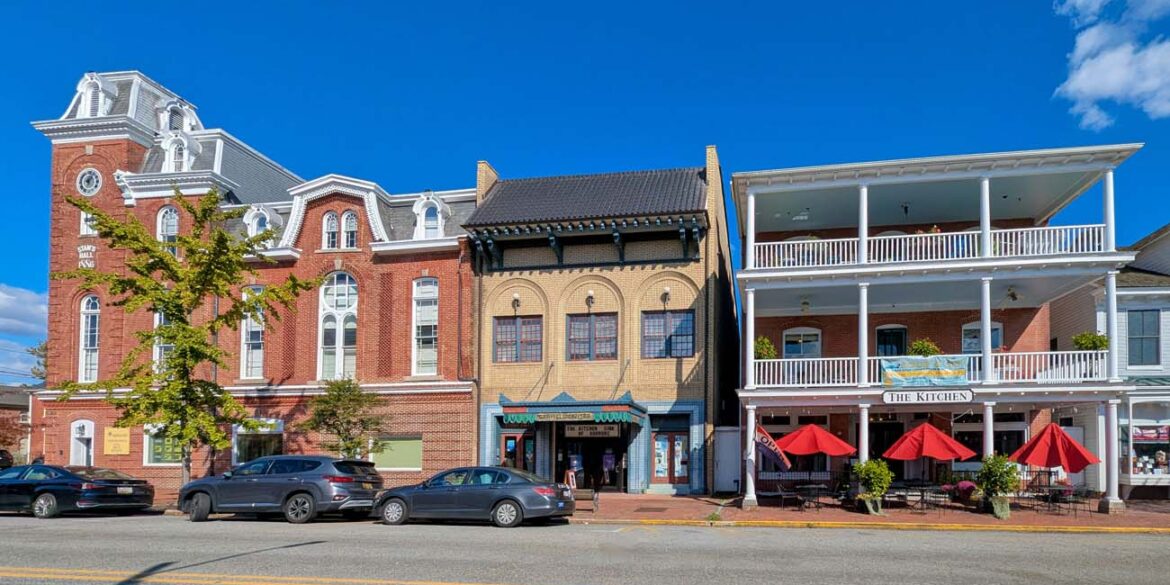 Historic downtown Chestertown Maryland street view showing row of preserved colonial and Victorian buildings including red brick Second Empire style town hall with clock tower, beige commercial building, and white building with double porches housing The Kitchen restaurant with red umbrellas, cars parked along street