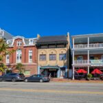 Historic downtown Chestertown Maryland street view showing row of preserved colonial and Victorian buildings including red brick Second Empire style town hall with clock tower, beige commercial building, and white building with double porches housing The Kitchen restaurant with red umbrellas, cars parked along street