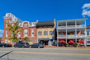Historic downtown Chestertown Maryland street view showing row of preserved colonial and Victorian buildings including red brick Second Empire style town hall with clock tower, beige commercial building, and white building with double porches housing The Kitchen restaurant with red umbrellas, cars parked along street