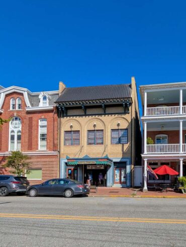 Historic downtown Chestertown Maryland street view showing row of preserved colonial and Victorian buildings including red brick Second Empire style town hall with clock tower, beige commercial building, and white building with double porches housing The Kitchen restaurant with red umbrellas, cars parked along street