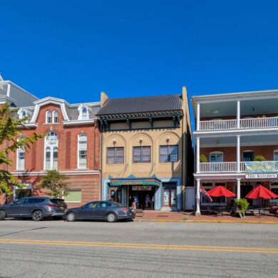 Historic downtown Chestertown Maryland street view showing row of preserved colonial and Victorian buildings including red brick Second Empire style town hall with clock tower, beige commercial building, and white building with double porches housing The Kitchen restaurant with red umbrellas, cars parked along street