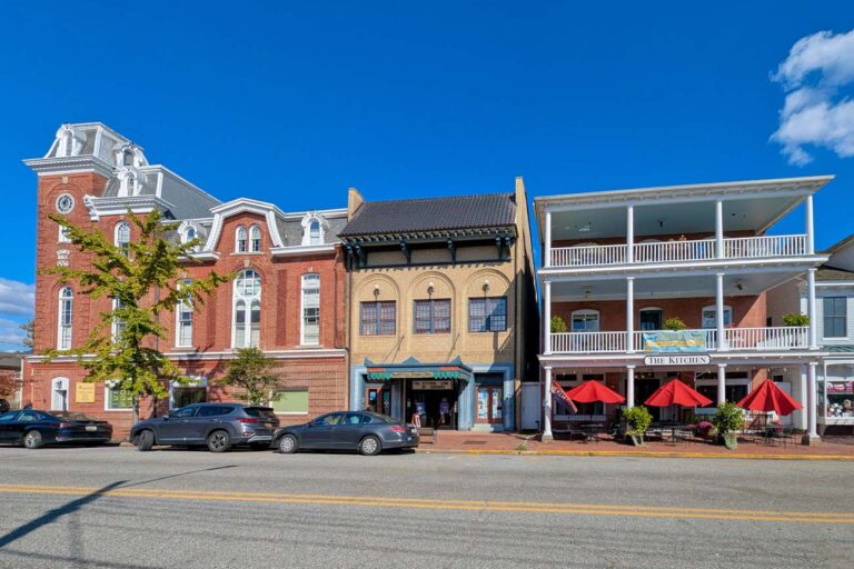 Historic downtown Chestertown Maryland street view showing row of preserved colonial and Victorian buildings including red brick Second Empire style town hall with clock tower, beige commercial building, and white building with double porches housing The Kitchen restaurant with red umbrellas, cars parked along street