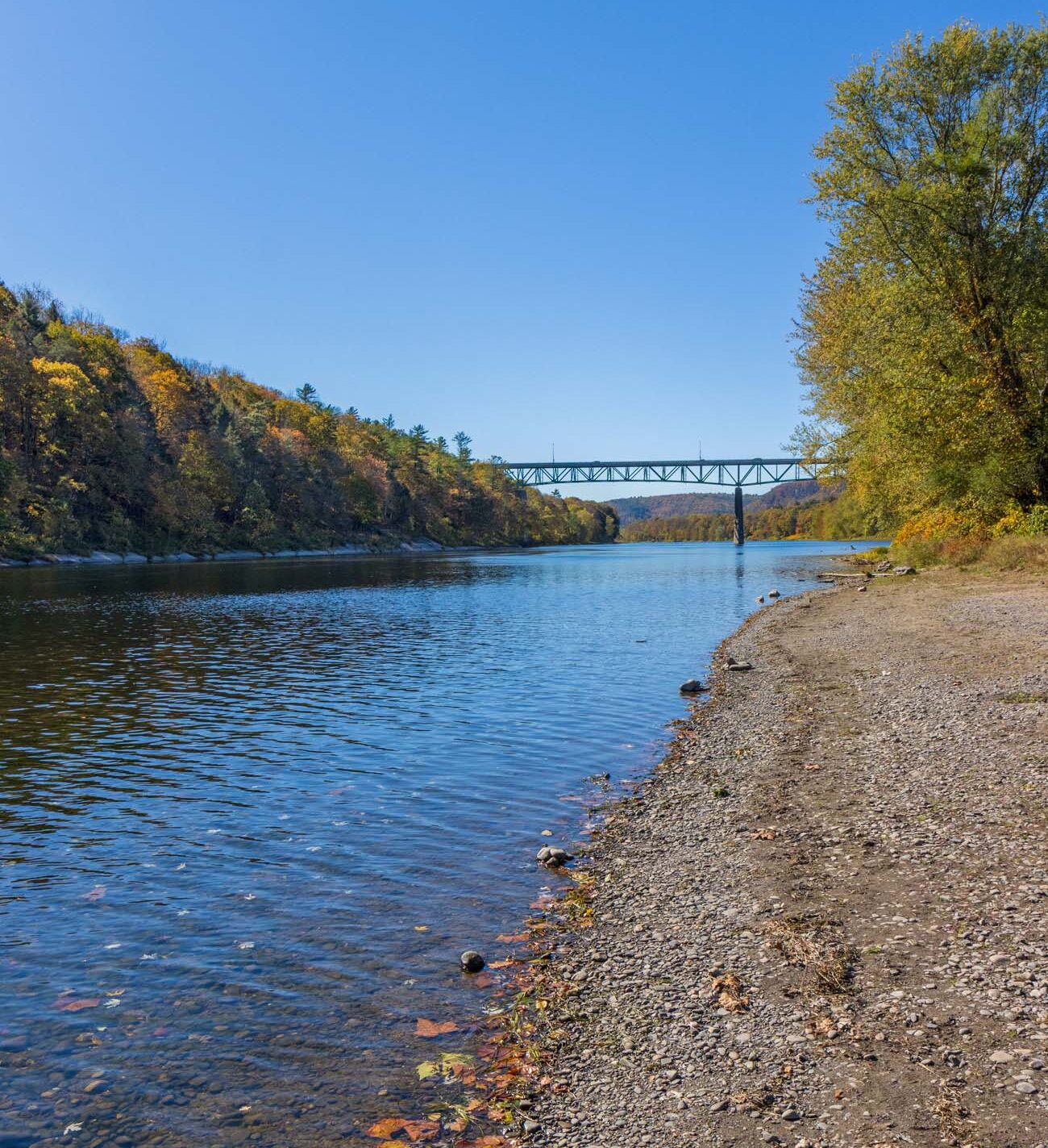 Scenic Delaware River shoreline at Milford Beach in autumn showing peaceful sandy beach, calm blue water, forested hillsides with fall foliage colors and pedestrian bridge, part of Delaware Water Gap National Recreation Area Pennsylvania