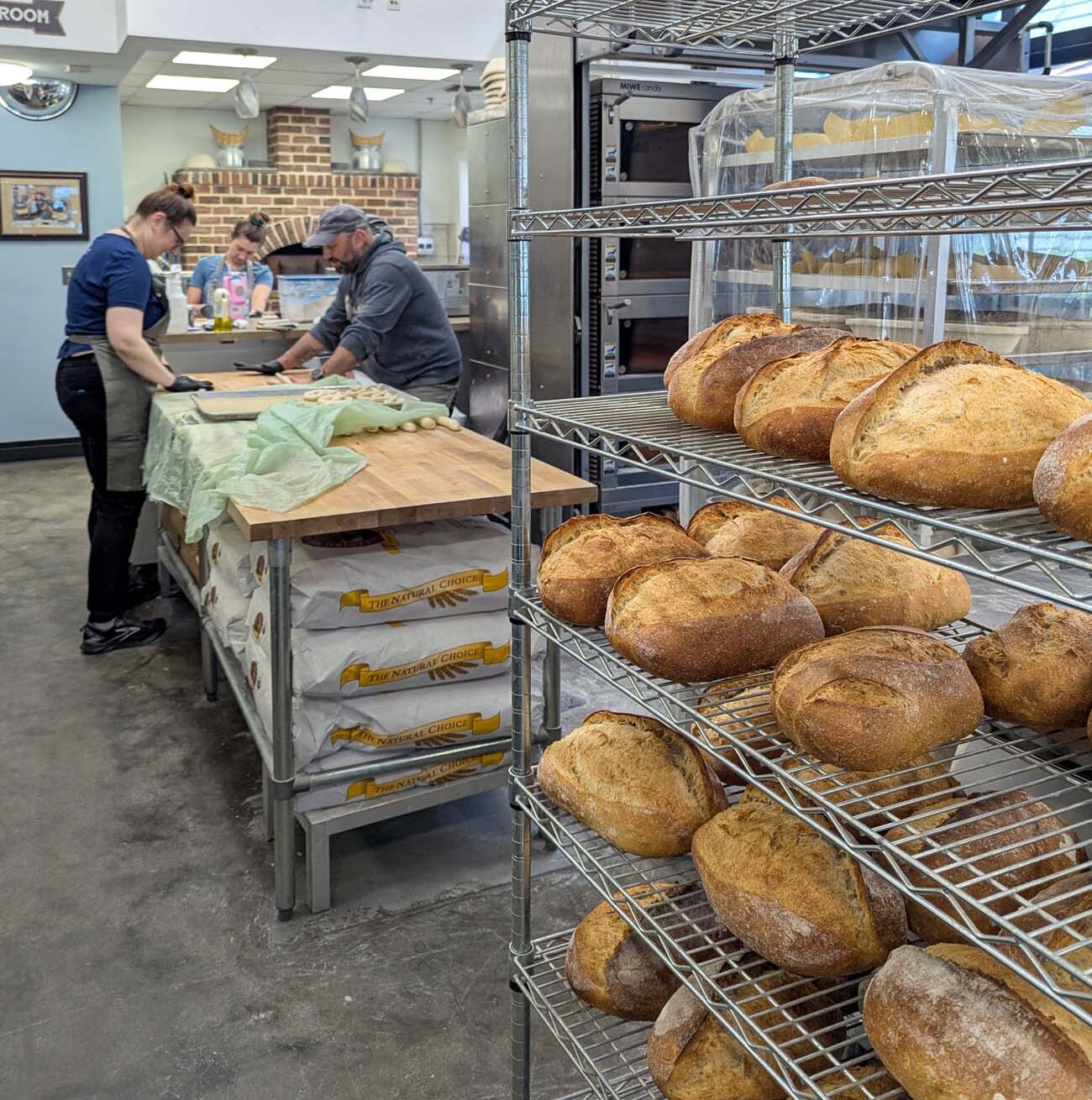 Things to Do in Chestertown MD: Eastern Shore Travel Guide 28 Behind-the-scenes view of Modern Stoneage Kitchen bakery showing professional bakers working at a large wooden table preparing dough. Wire shelving racks in the foreground display dozens of freshly baked artisan bread loaves with golden-brown crusts dusted with flour. A brick oven is visible in the background along with stainless steel commercial baking equipment. The industrial kitchen features polished concrete floors and bright overhead lighting.
