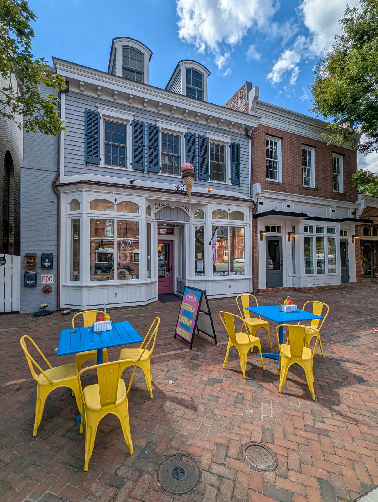 Things to Do in Chestertown MD: Eastern Shore Travel Guide 18 Stam's Luncheonette exterior showing historic white two-story building with black shutters, curved bay windows on first floor, red door entrance, bright yellow metal bistro tables and chairs on brick plaza, ice cream cone sign, blue sky
