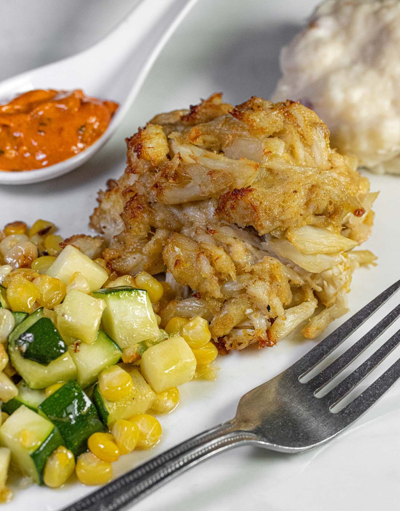 Things to Do in Chestertown MD: Eastern Shore Travel Guide 20 lose-up of a golden-brown jumbo lump crab cake with crispy edges on a white plate, accompanied by a side of sautéed zucchini and corn succotash. A ramekin of orange remoulade sauce and what appears to be coleslaw are visible in the background. A silver fork rests on the right side of the plate. The crab cake shows large chunks of premium crab meat with minimal filler and a perfectly seared crust.