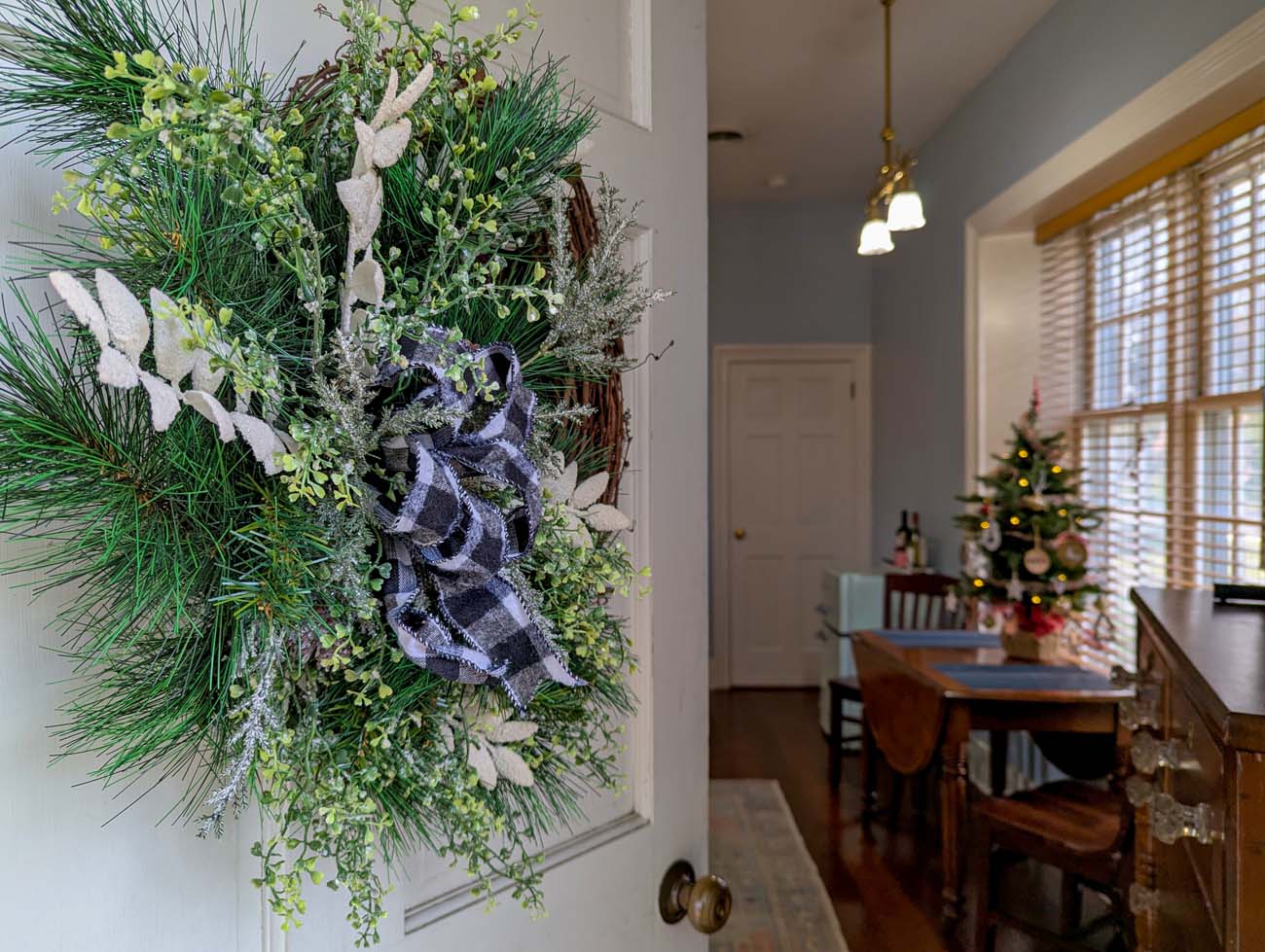 Elegant wreath with buffalo check ribbon, white frosted branches, and eucalyptus hanging on white door, Christmas tree visible through doorway in formal dining room beyond, holiday home decor