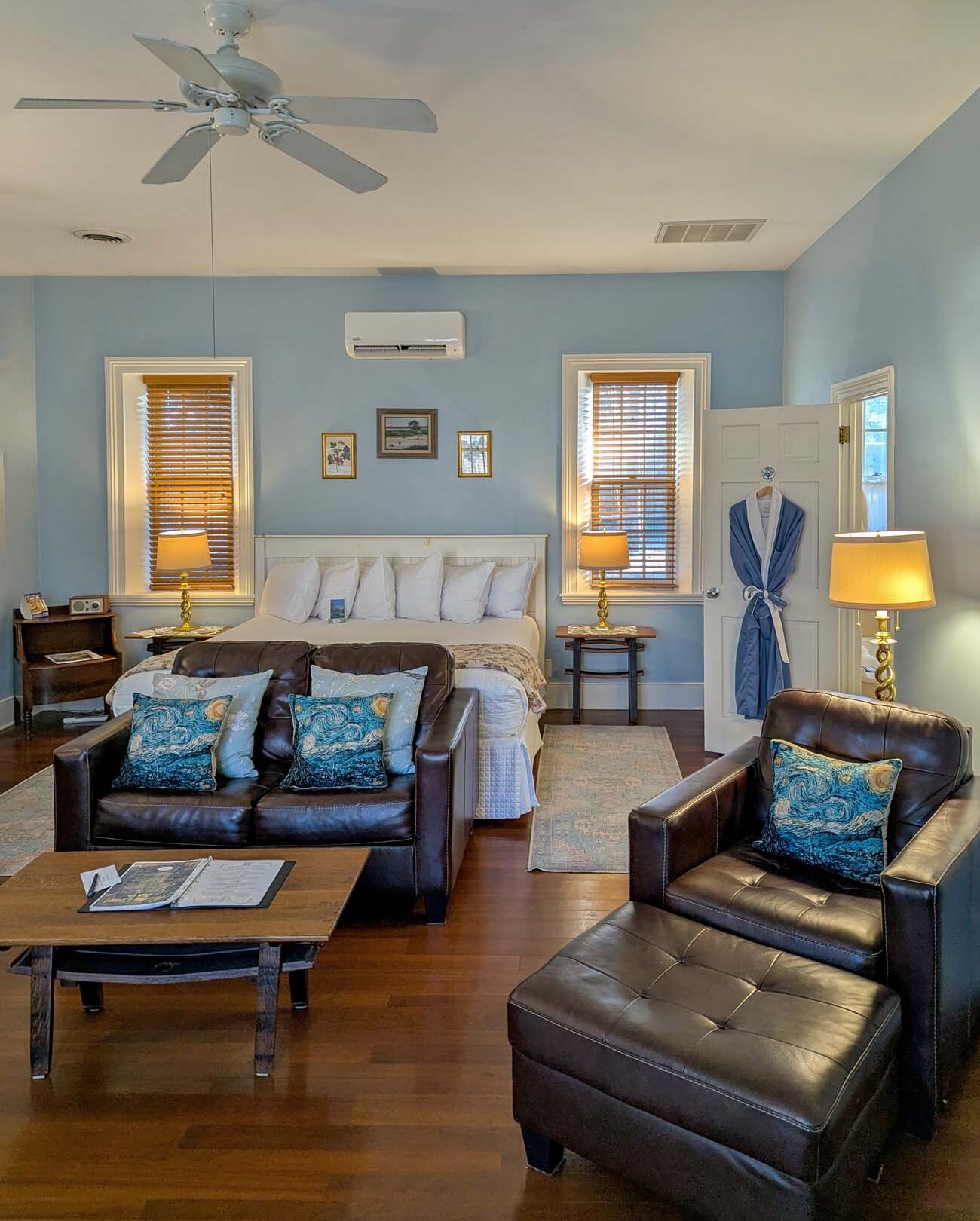 Sunrise room at Brampton Garden Cottage showing king bed with white linens, brown leather loveseat and chair, blue walls, ceiling fan, air conditioning unit, warm lamps, hardwood floors, bathroom door visible, spacious 475 square foot accommodation