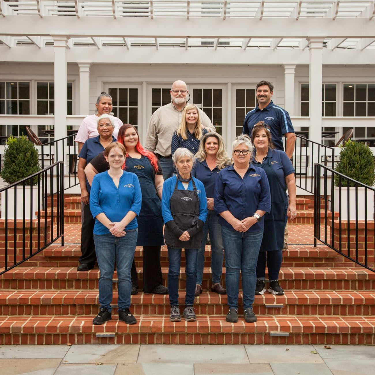 Brampton 1860 staff and owners posing on brick front steps of white-columned Manor House porch, group of 11 people in navy blue Brampton polo shirts and casual attire, smiling team photo showing diverse staff members