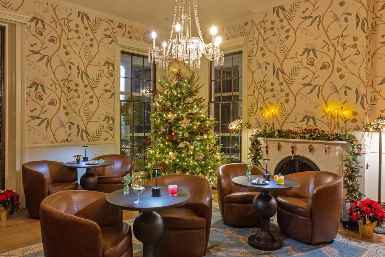 Brampton 1860 Salon de Lumiere decorated for Christmas with tall lit tree adorned with white poinsettias and red ornaments under crystal chandelier, brown leather club chairs at small round tables with festive cocktails, elegant botanical wallpaper, fireplace with evergreen garland and red ornaments, Tiffany-style lamps, warm ambient lighting in newly renovated gathering space