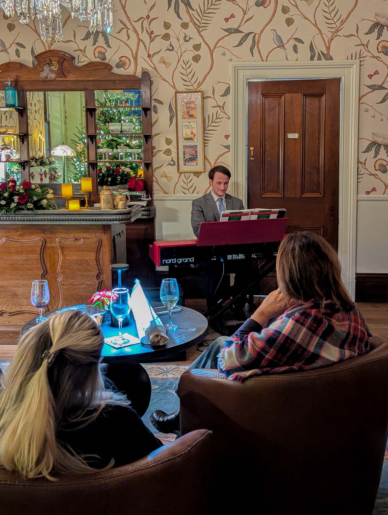 Michael Casey playing red keyboard piano in Brampton's Salon de Lumiere during holiday social hour, guests seated in foreground watching performance, ornate bar with mirror and Christmas decorations in background, chandelier overhead, elegant patterned wallpaper