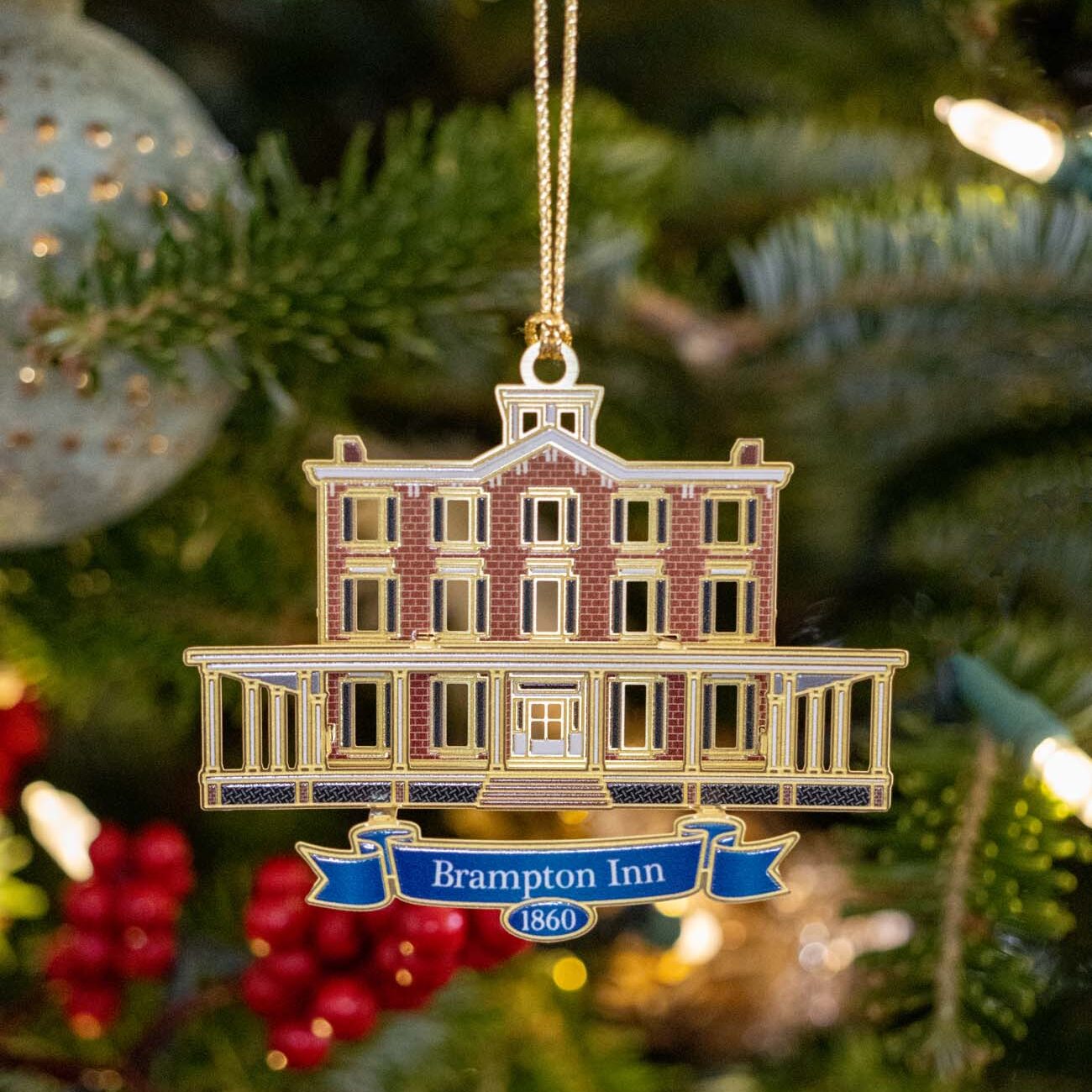 Custom Brampton Inn 1860 Christmas ornament showing detailed illustration of three-story brick manor house with columned porch, blue ribbon banner, hanging on evergreen tree with red berry clusters and bokeh lights