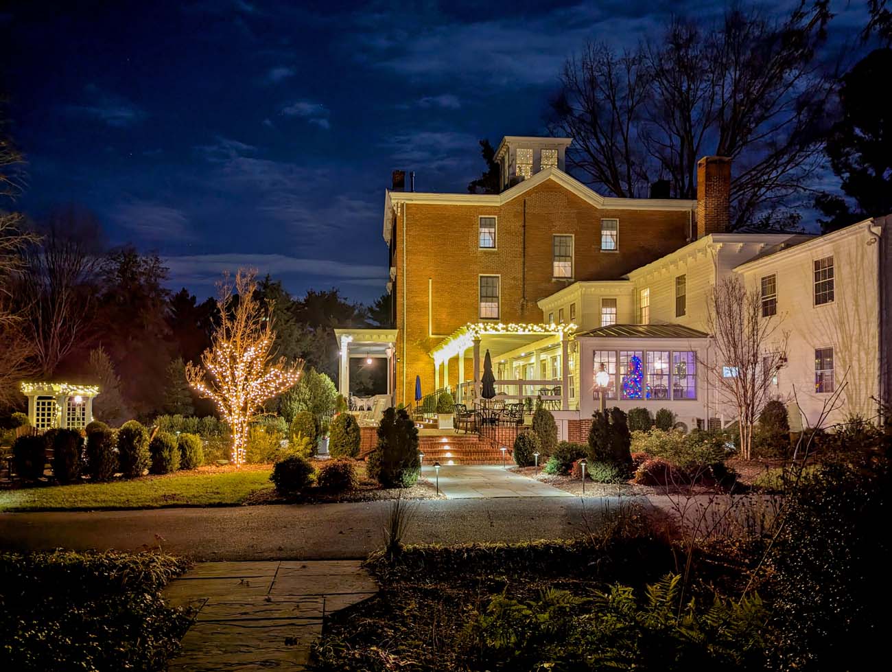 Brampton 1860 Manor House illuminated at night during holidays with white string lights outlining porch columns and roofline, Christmas tree visible in lit window, bare winter trees silhouetted against dark blue twilight sky, brick walkway and landscaped grounds