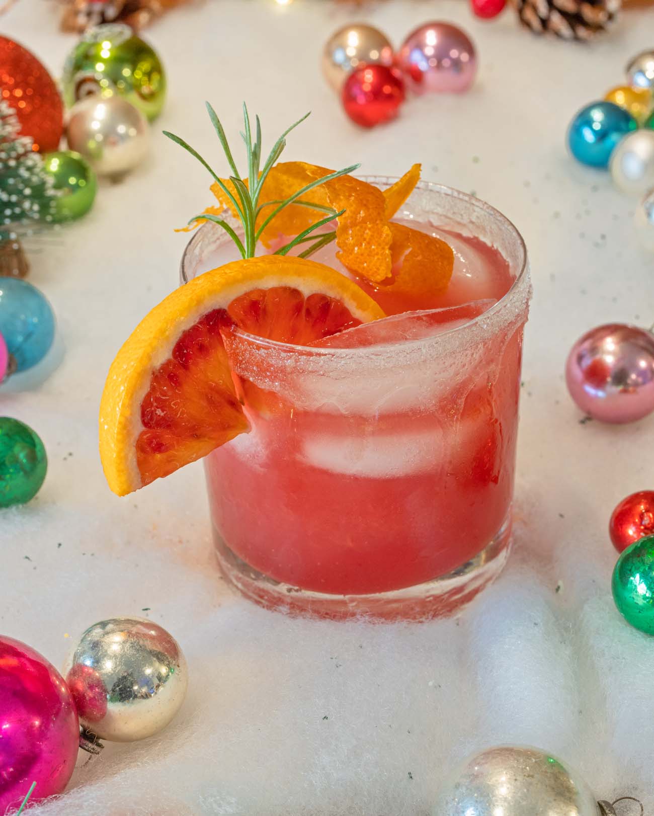 Pink grapefruit cocktail in sugar-rimmed rocks glass garnished with blood orange wheel, rosemary sprig, and candied orange peel, surrounded by vintage Christmas ornaments on white surface
