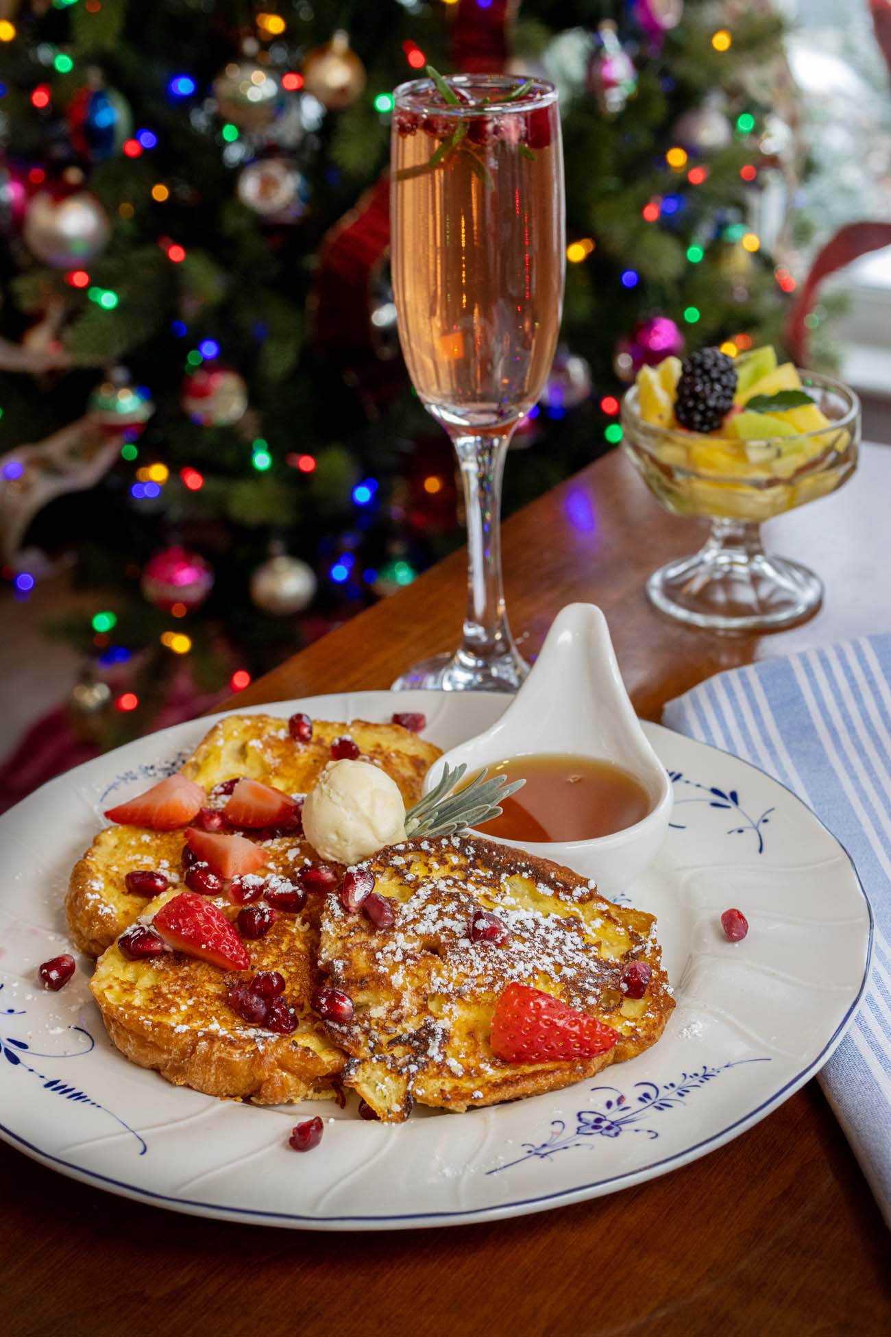 Holiday French toast breakfast plate with pomegranate seeds and fresh strawberries, served with champagne flute, fruit compote in glass dish, Christmas tree with multicolored lights in background, white china with blue floral pattern