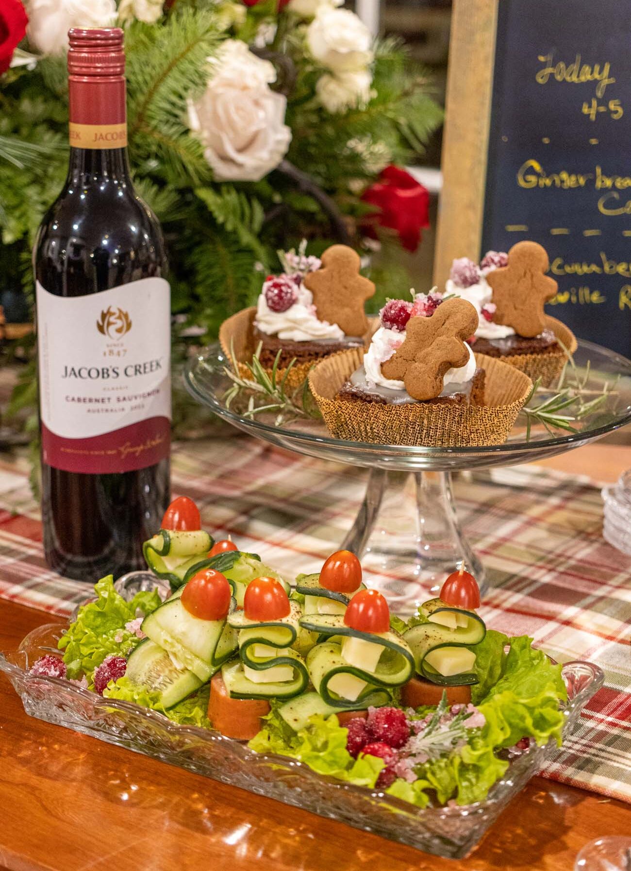 Holiday tea time spread showing Christmas tree-shaped sugar cookies with green frosting and sprinkles, gingerbread cupcakes with whipped cream and gingerbread men, vegetable crudité platter with cherry tomatoes and cucumber ribbons, Jacob's Creek wine bottle, festive presentation on holiday plaid tablecloth