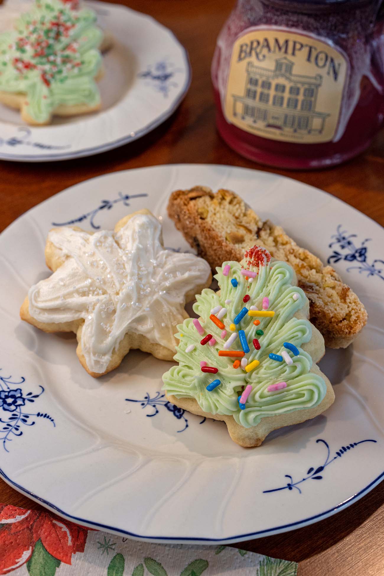 Assorted holiday cookies on decorative china plate including frosted Christmas tree sugar cookie with sprinkles, plain snowflake cookie, winter biscotti, served with Brampton-branded beverage in background